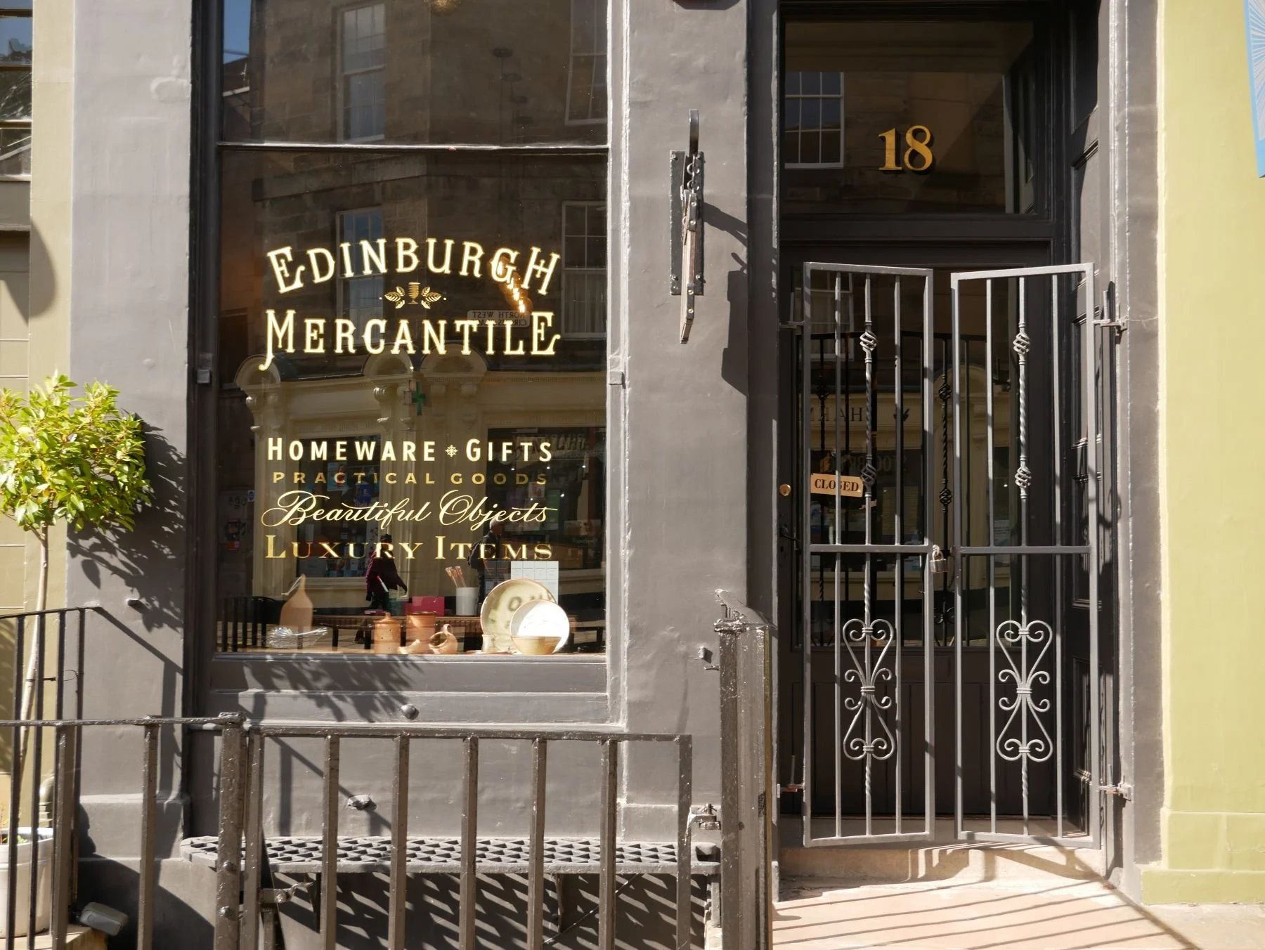 The storefront of Edinburgh Mercantile, a homeware, gifts, and luxury items store, with a large glass window displaying ceramics and decorative objects inside. The store has a black metal gate and door, and a small green bush on the sidewalk.