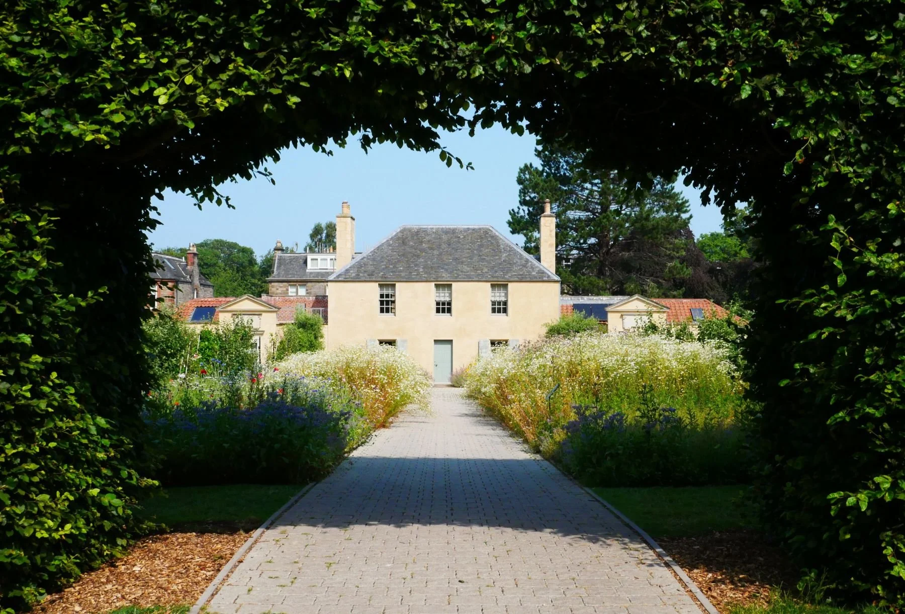 View of a building in Edinburgh's Botanic Gardens seen through a garden archway with a stone pathway, surrounded by flowering plants and trees.