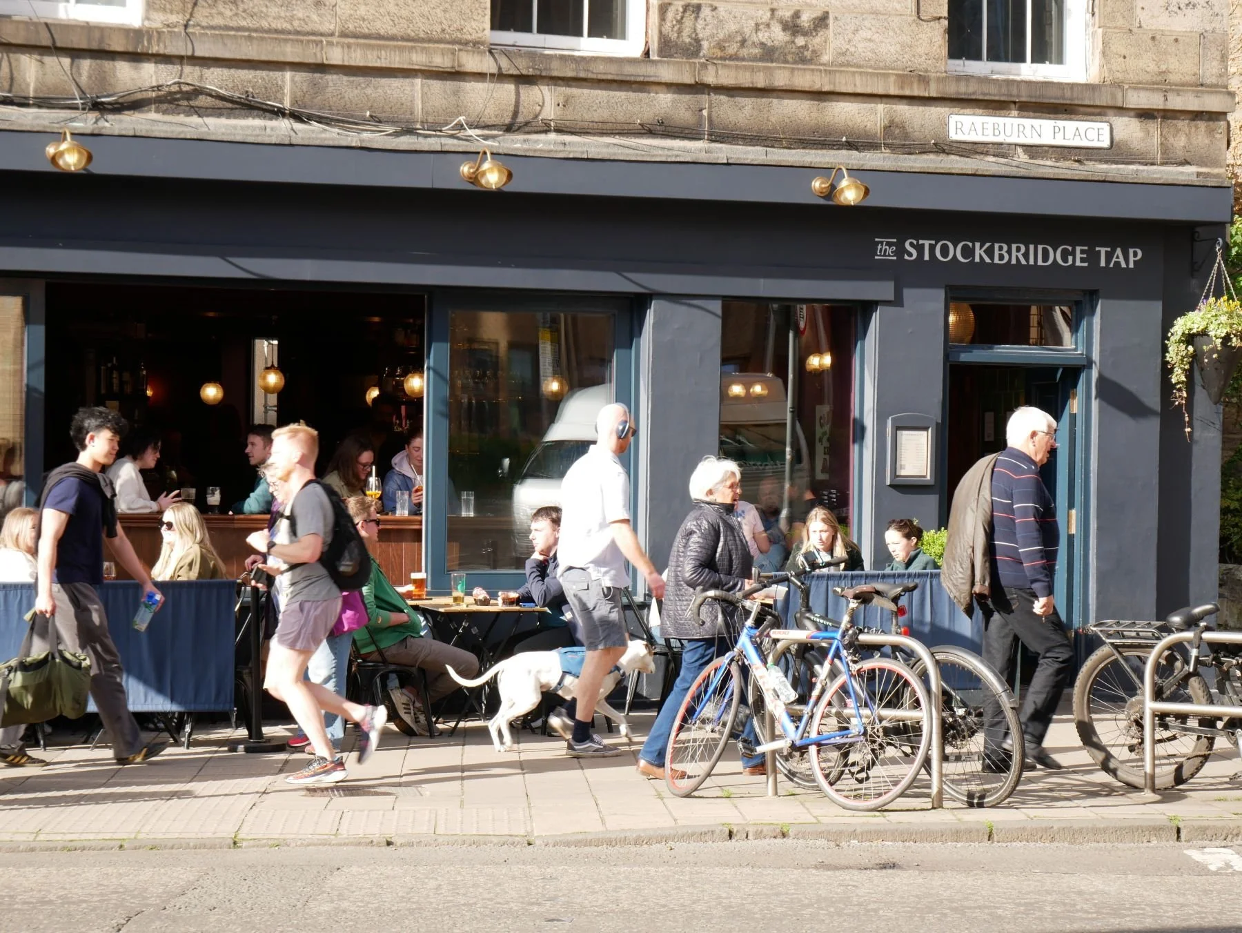 People sitting and standing outside a cafe called The Stockbridge Tap, with bicycles parked along the sidewalk, and some individuals walking, chatting, and using their phones on a sunny day.