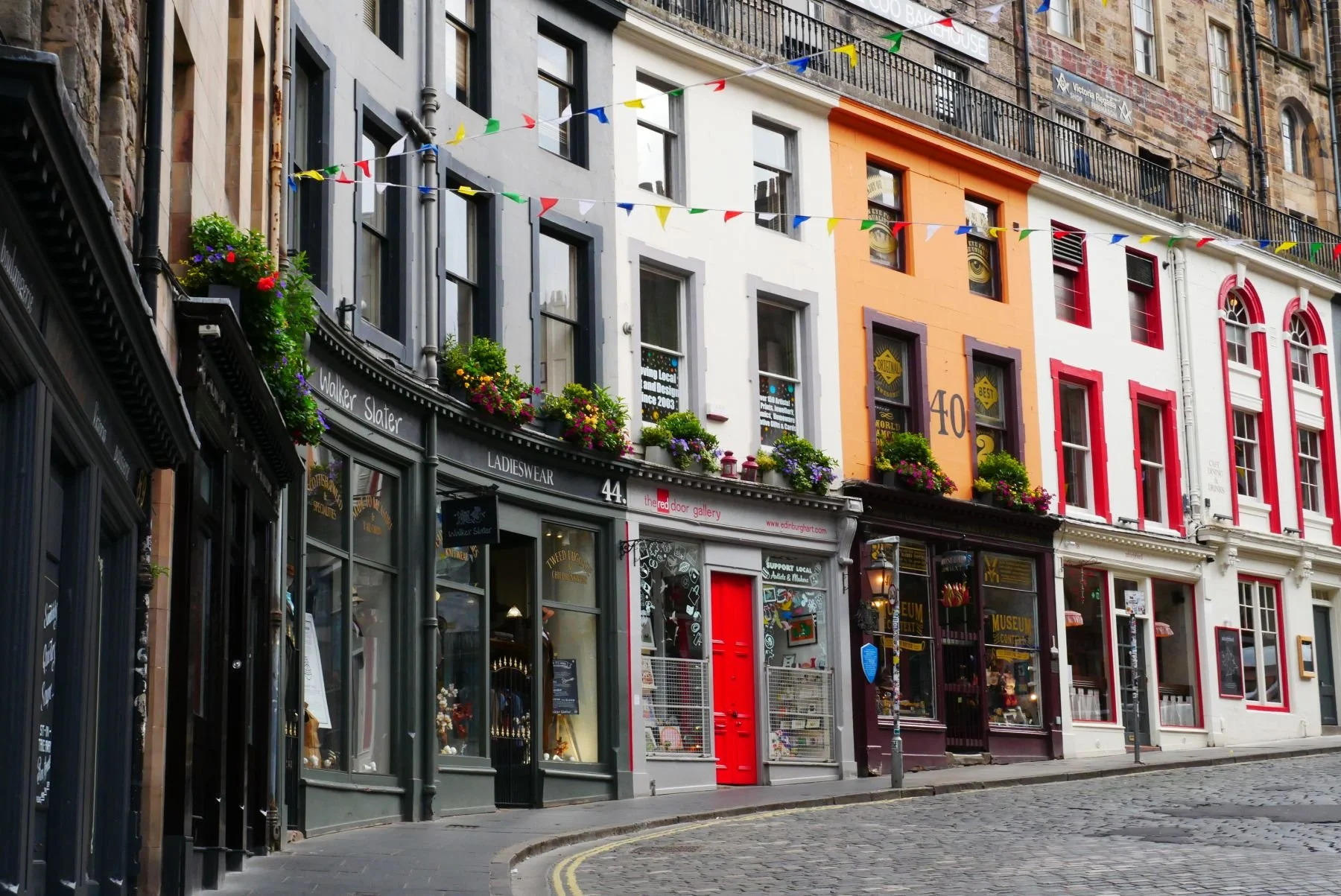 Colorful buildings on Victoria Street, Edinburgh, with shops storefronts and flower boxes on a cobblestone street, decorated with string lights.
