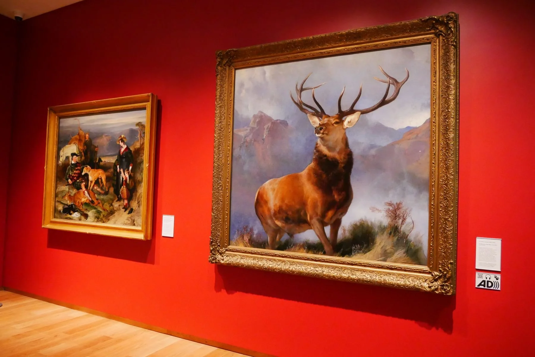 Display a painting in an art gallery with red walls. The painting features a large with impressive antlers, standing on a mountainous Highland landscape.