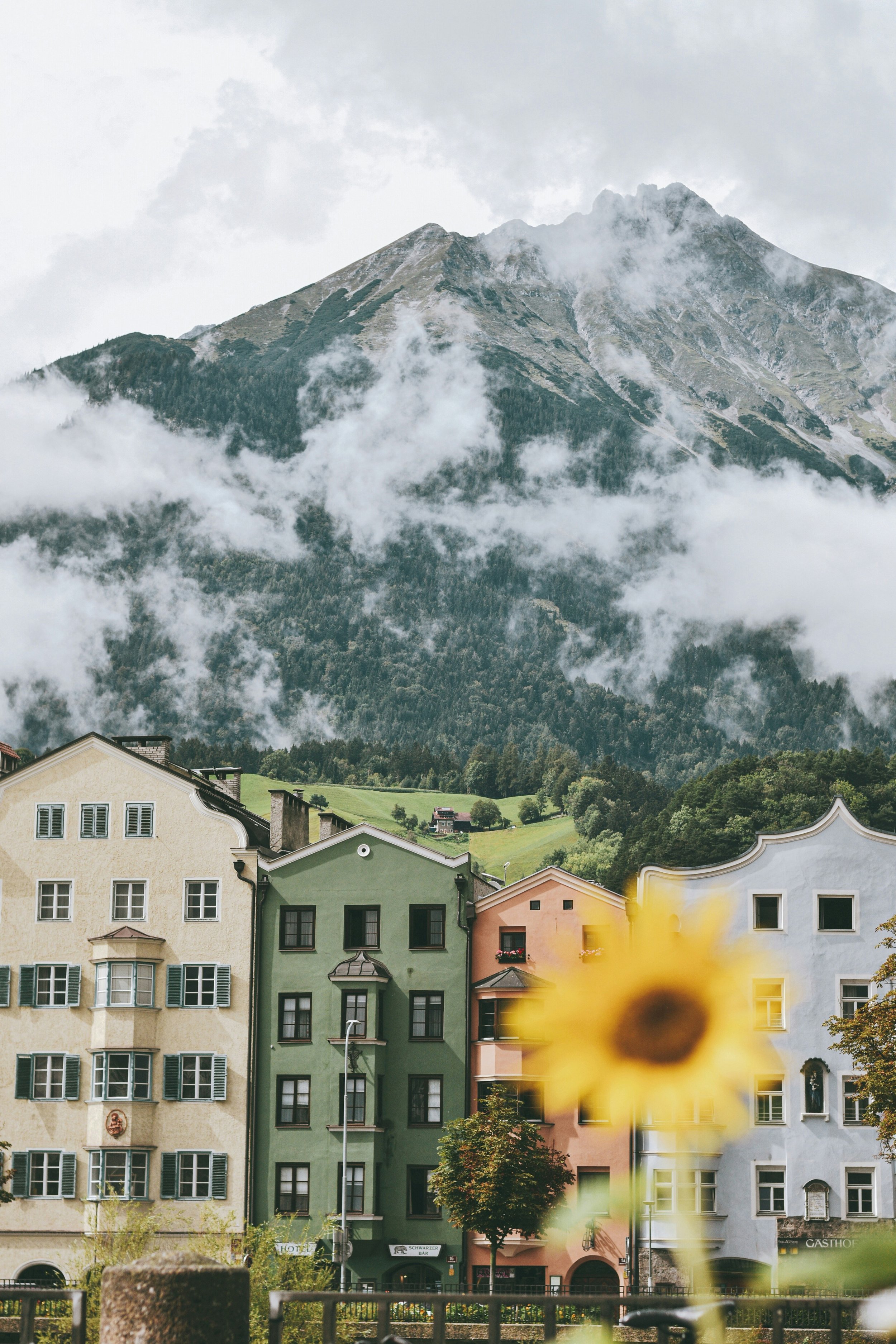 Colorful multi-story buildings with arched and rectangular windows, set against a mountainous backdrop with clouds and mist, in a European town.