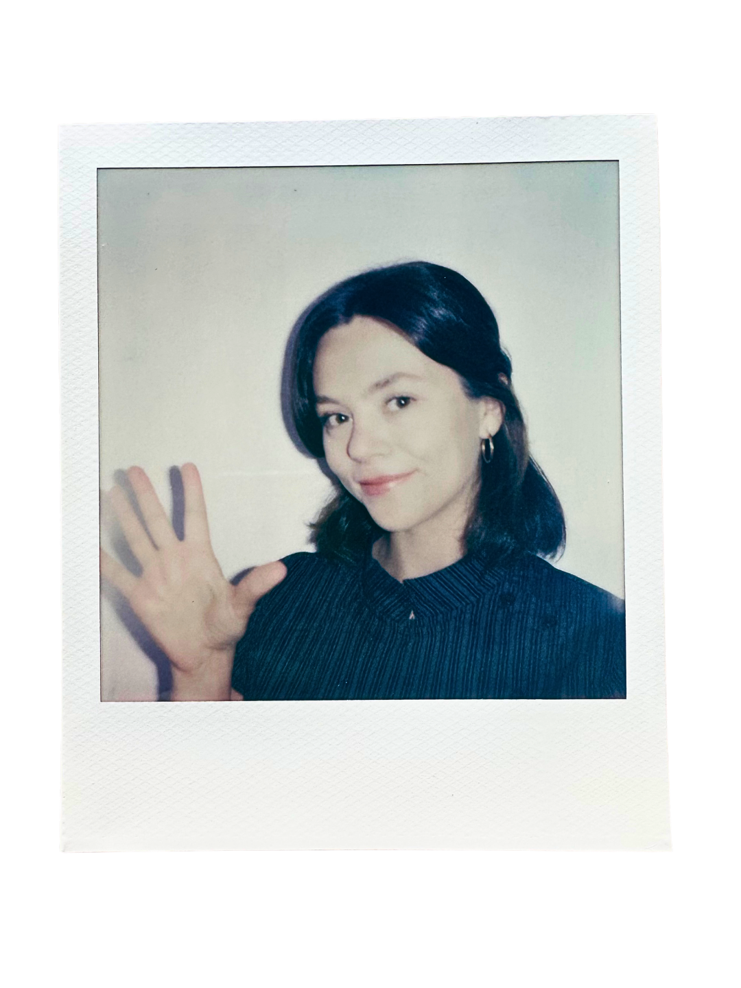A young woman with dark hair and hoop earrings, smiling and waving at the camera, wearing a dark striped shirt, against a plain light background.