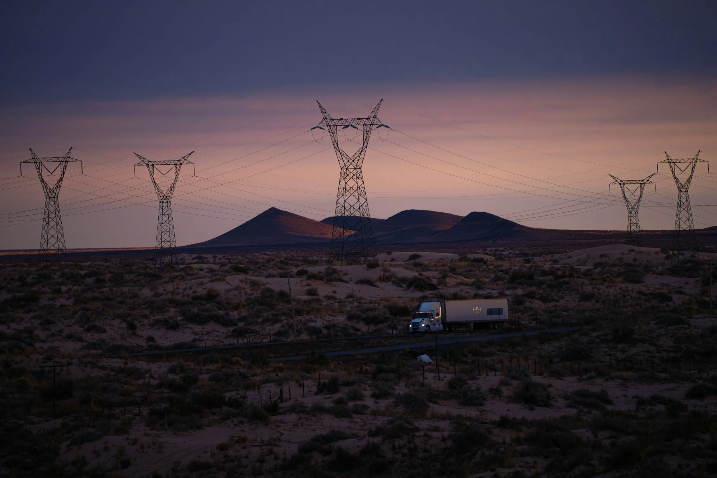 Electric power transmission towers in a desert landscape at dusk, with mountains in the background and a semi-truck parked on the road.