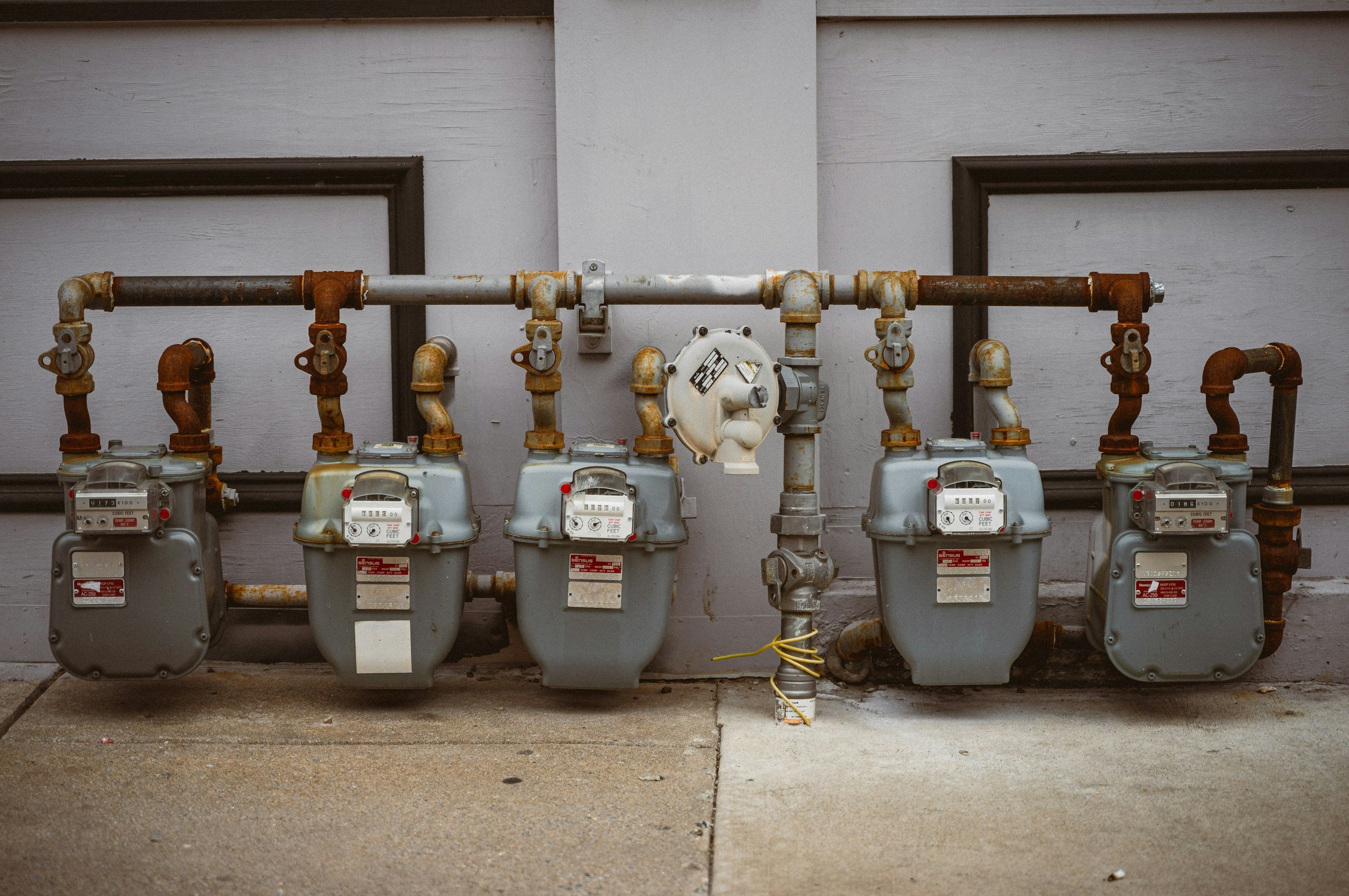 Multiple gas meters and rusted pipes attached to a white exterior wall of a building.