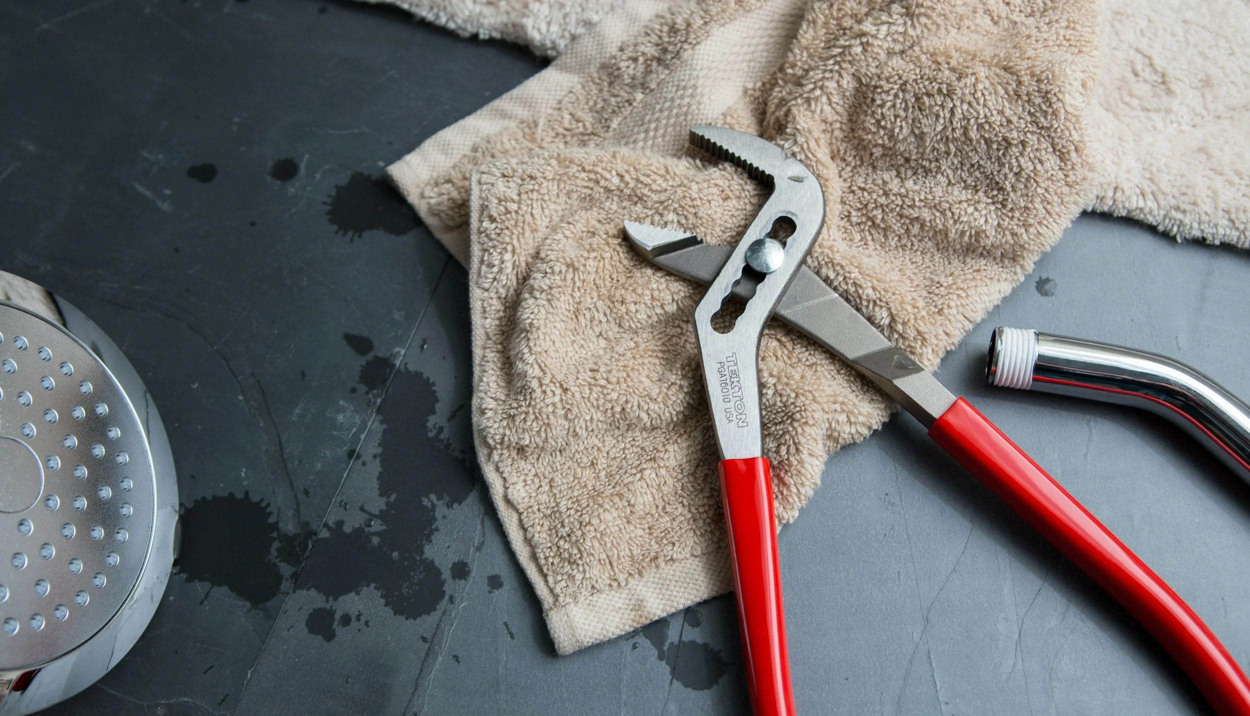 A soaked beige towel, a pair of red-handled water pliers, a metallic adjustable wrench, a metallic L-shaped pipe, and a metallic drain cover with small holes on a dark wet surface.