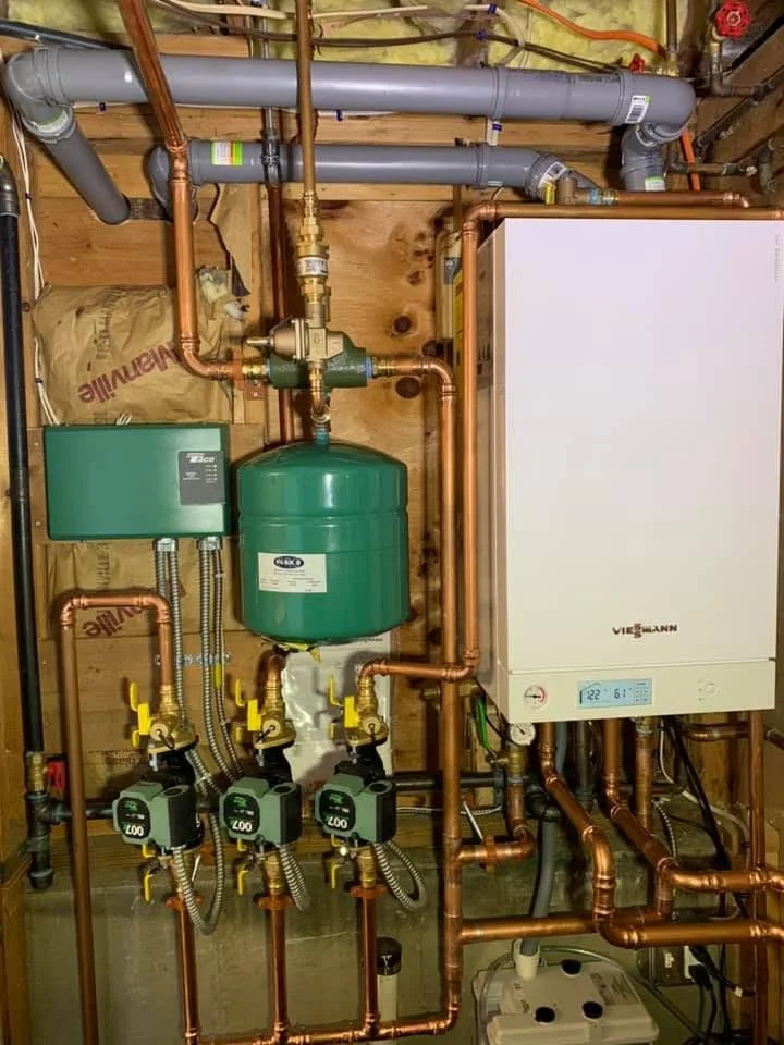 A plumbing setup with copper pipes, gray PVC pipes, a green expansion tank, a white boiler, and yellow shut-off valves mounted on a wooden wall.