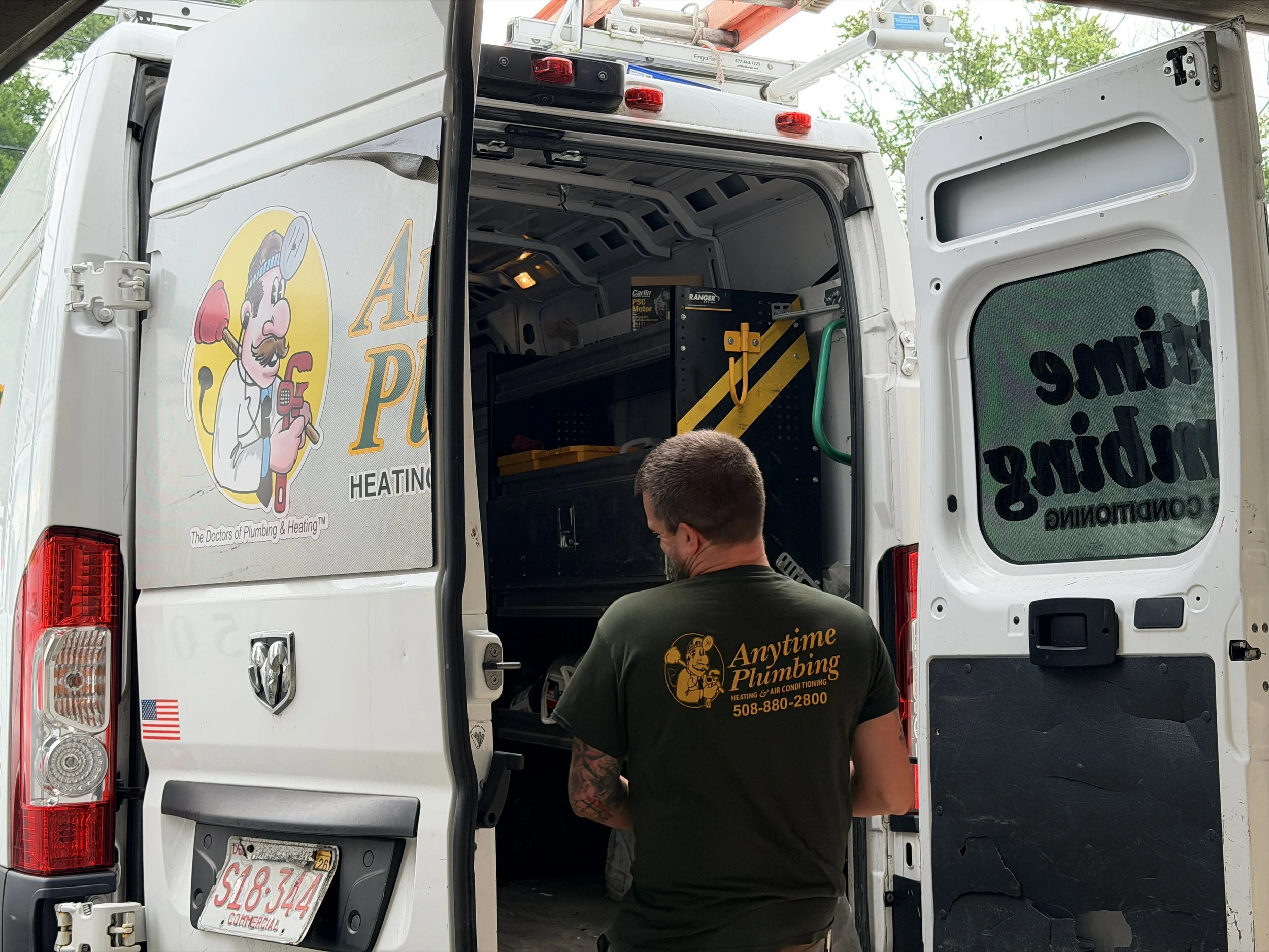 The back of a white service van with an open side door showing tools and equipment inside. A worker in a black shirt with gold and white text on the back is visible from behind, standing near the open door. The van's side features a cartoon logo of a