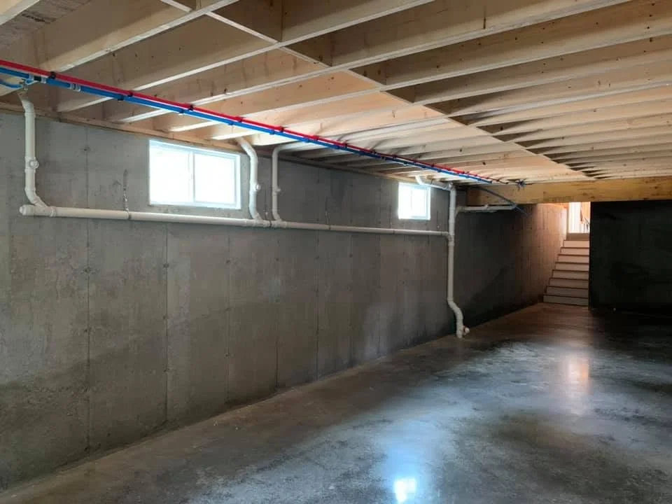 Underground basement with concrete walls, small windows, exposed pipes, wooden ceiling beams, and stairs leading to an upper level.
