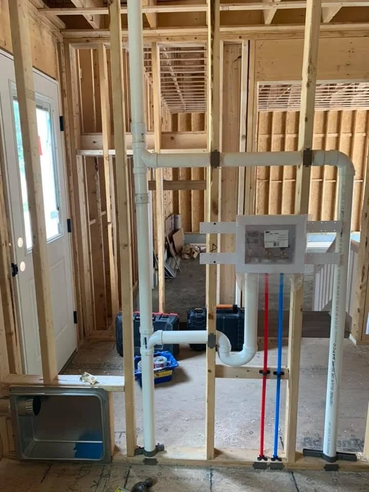 Interior of a house under construction with exposed wood framing, plumbing pipes, and electrical wiring.