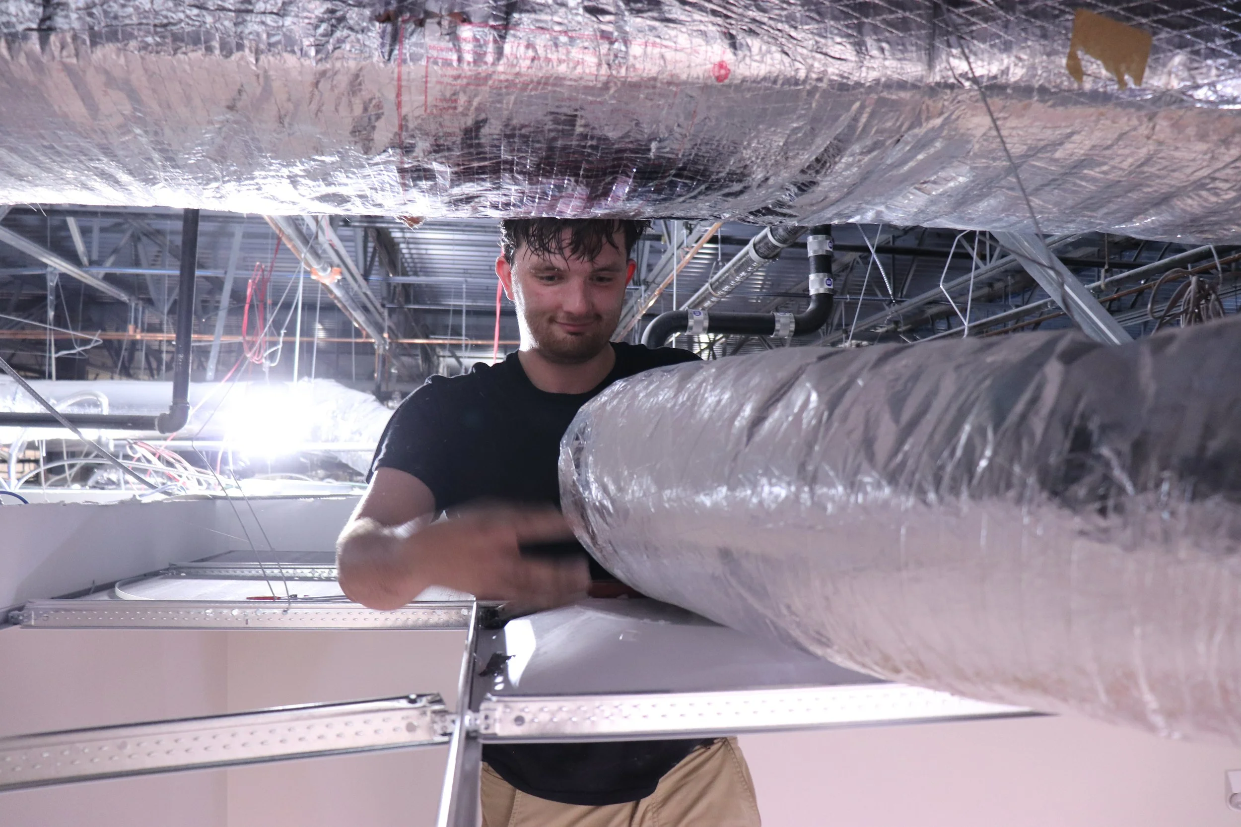 A young man with wet hair working underneath a large metallic pipe in an industrial or manufacturing setting, with exposed pipes and wiring above him.