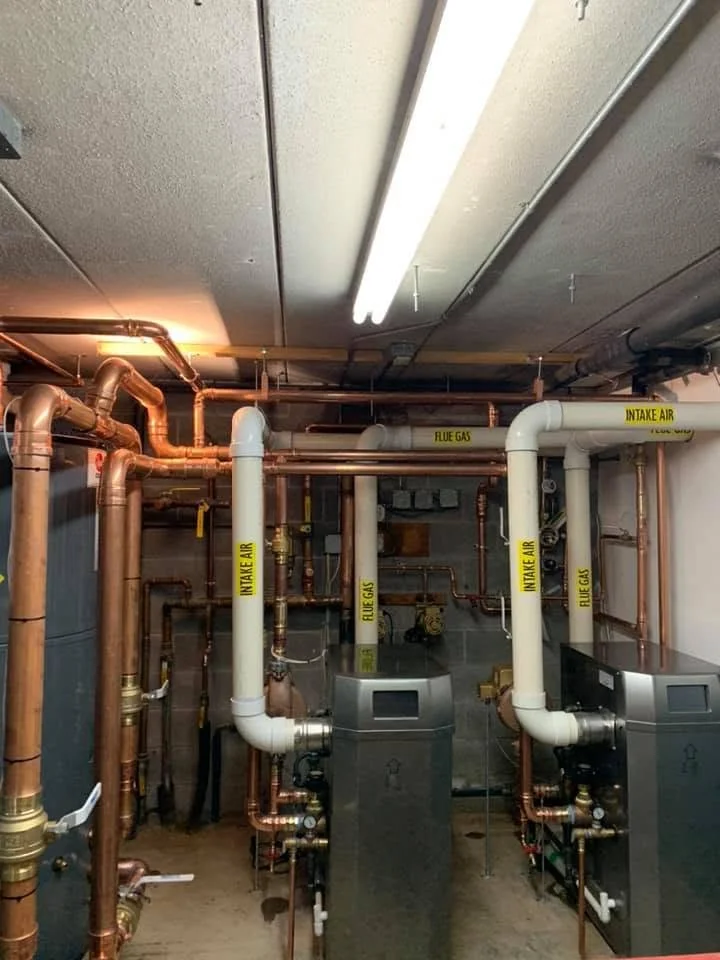 Industrial mechanical room with copper and steel pipes, two large gray machines, and yellow labels indicating intake air and flue gas.