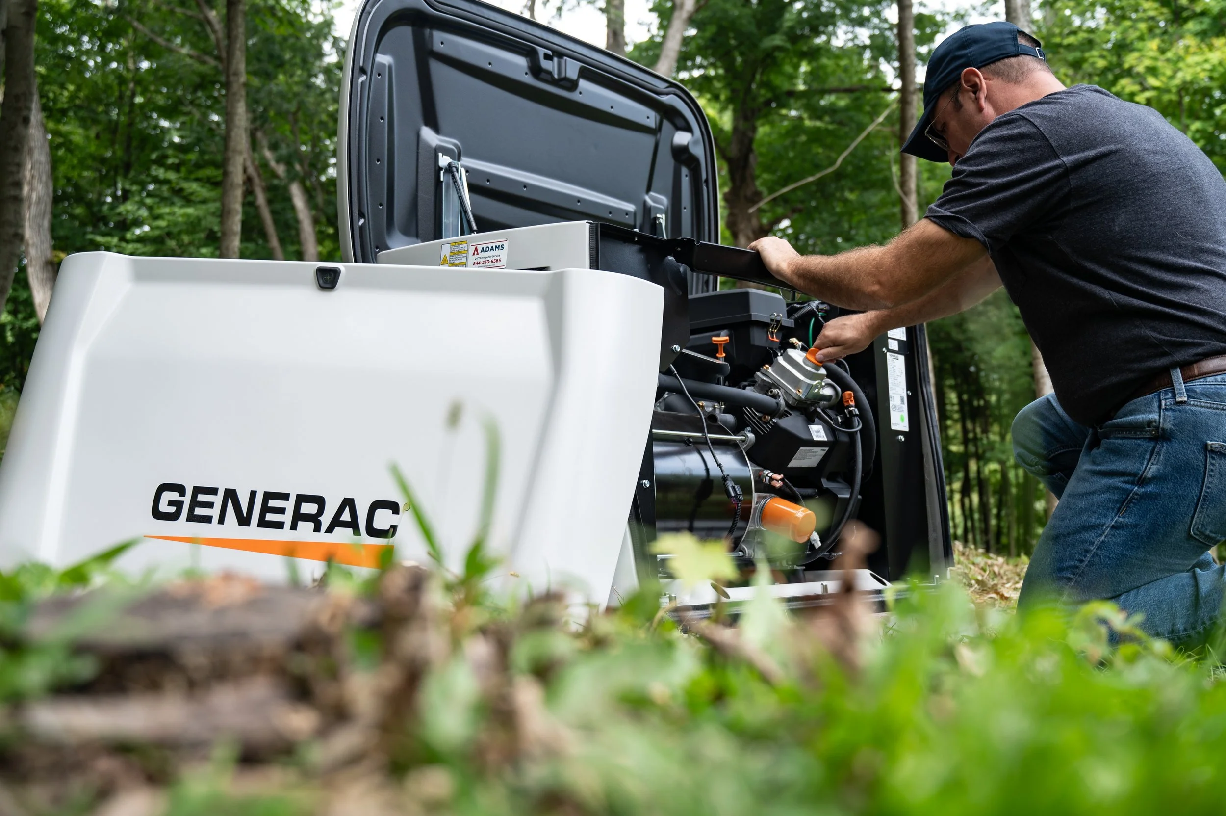 A man in a black shirt and cap working on a generator in a wooded outdoor area, with trees and green foliage in the background.