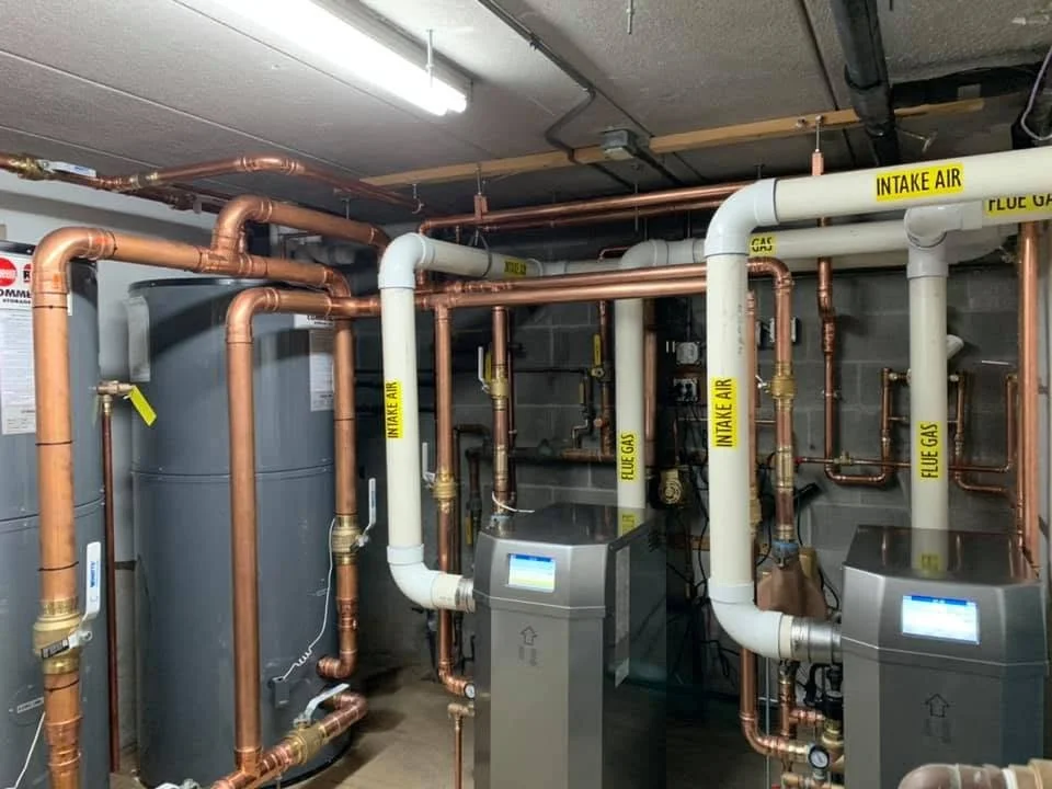 Mechanical room with copper and white PVC pipes labeled "INTAKE AIR," "FLUE GAS," and "FUME GAS," connected to large grey tanks and control systems.