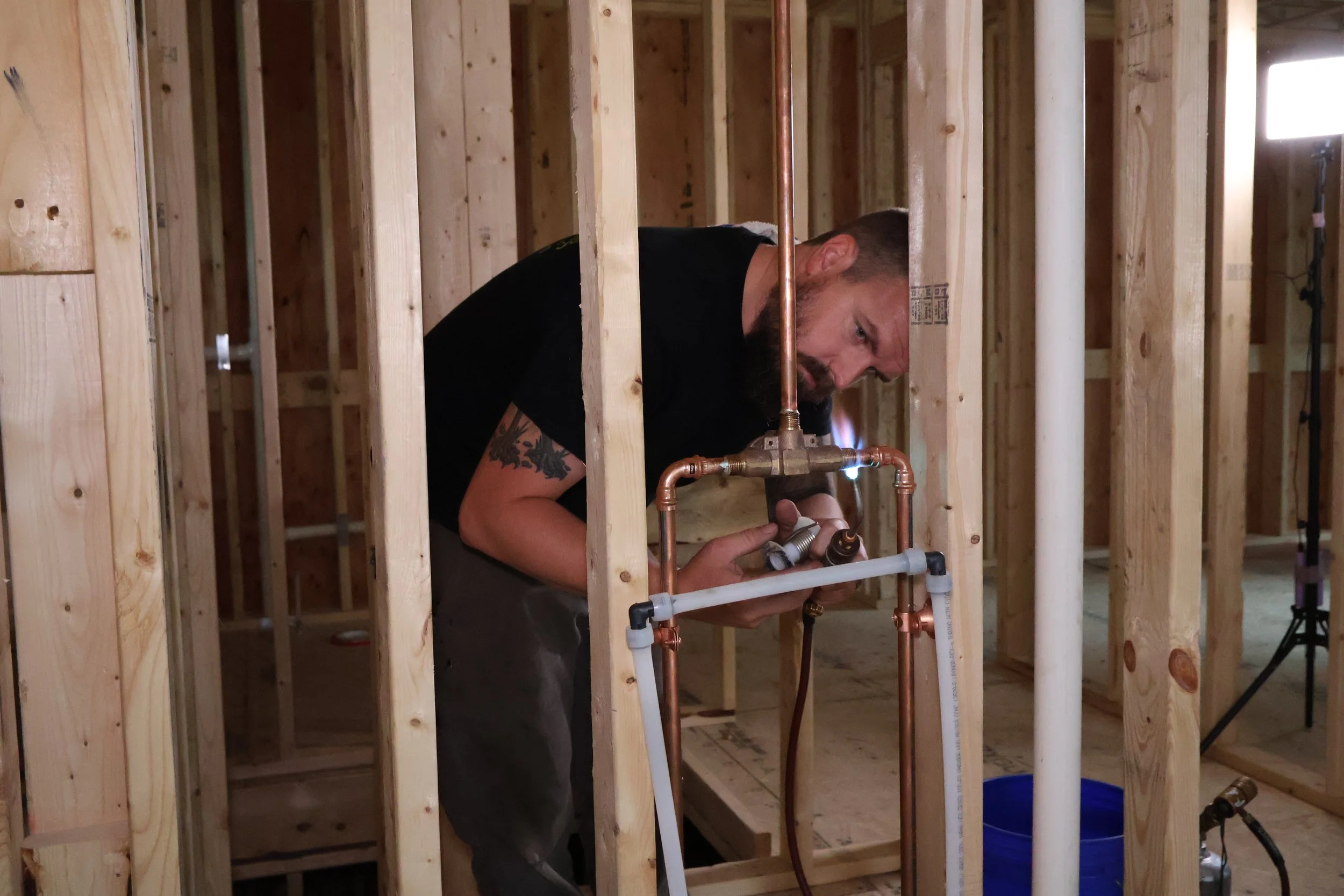 A man with a beard working on plumbing inside a wooden framed house, holding a pipe fitting and using a tool, with copper pipes and white PVC pipes around him.
