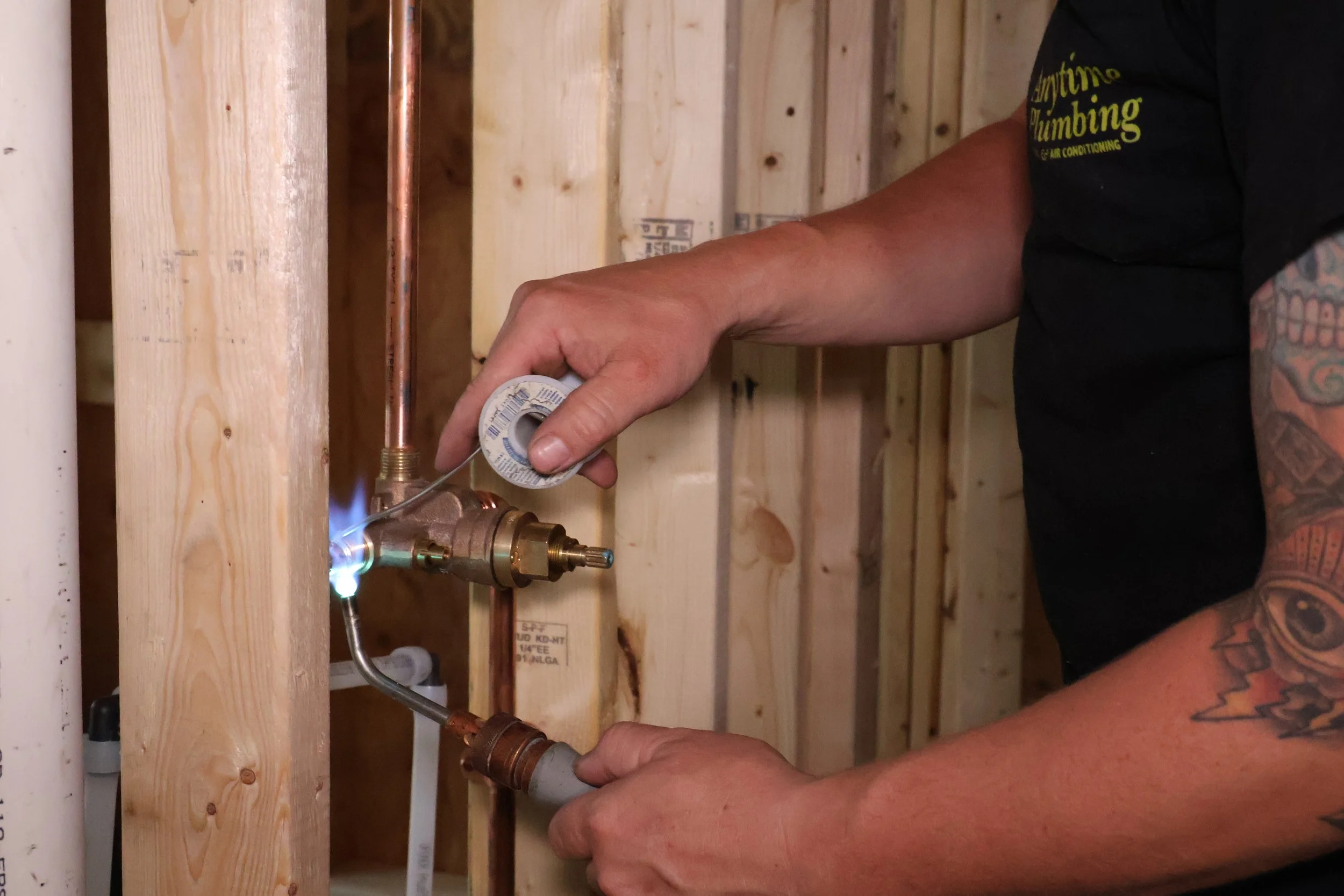 A person with tattoos on their arm installing a copper pipe and fitting in a wooden wall for plumbing.
