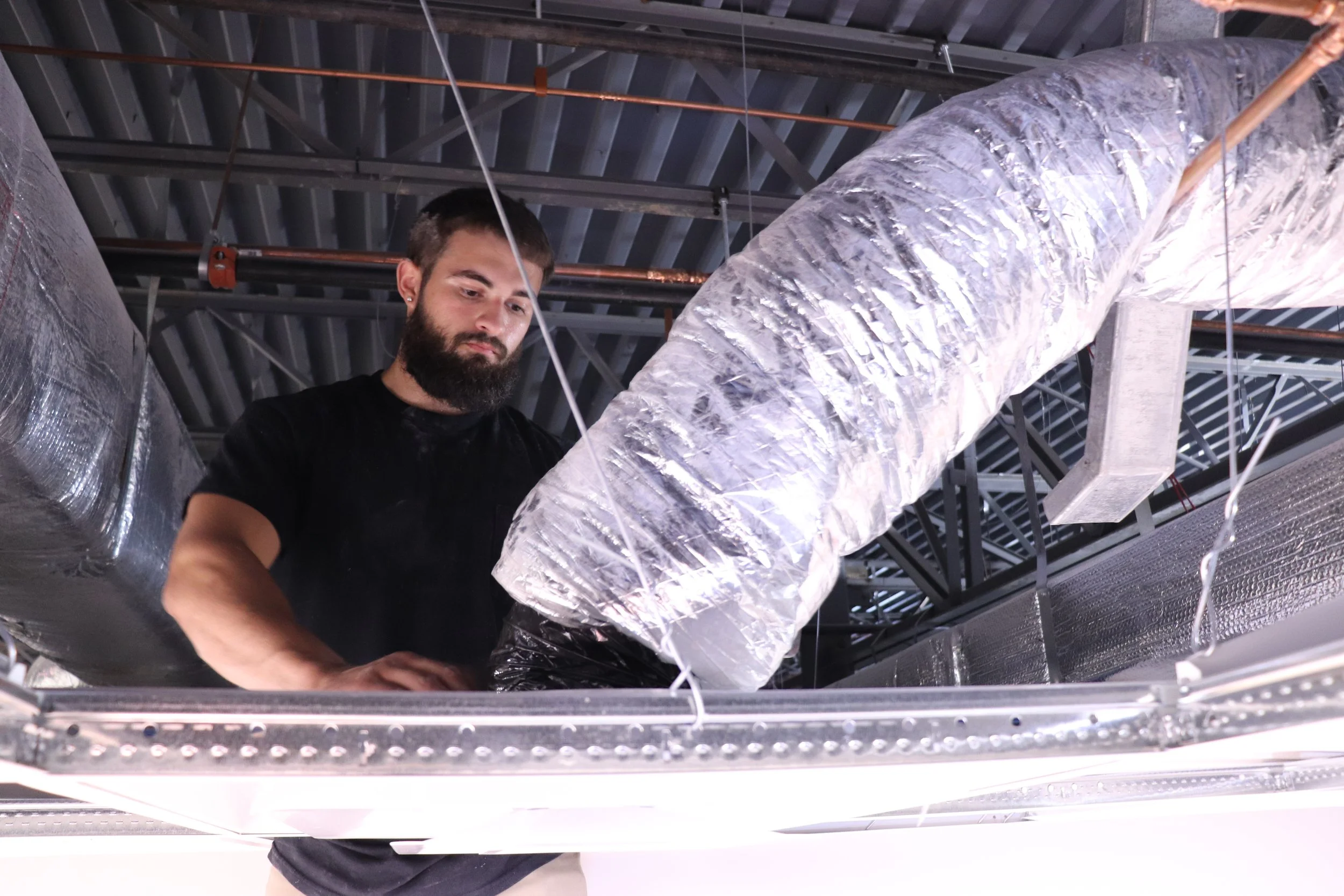 A man with a beard and earrings working in an industrial setting surrounded by large insulated pipes and metal framework.