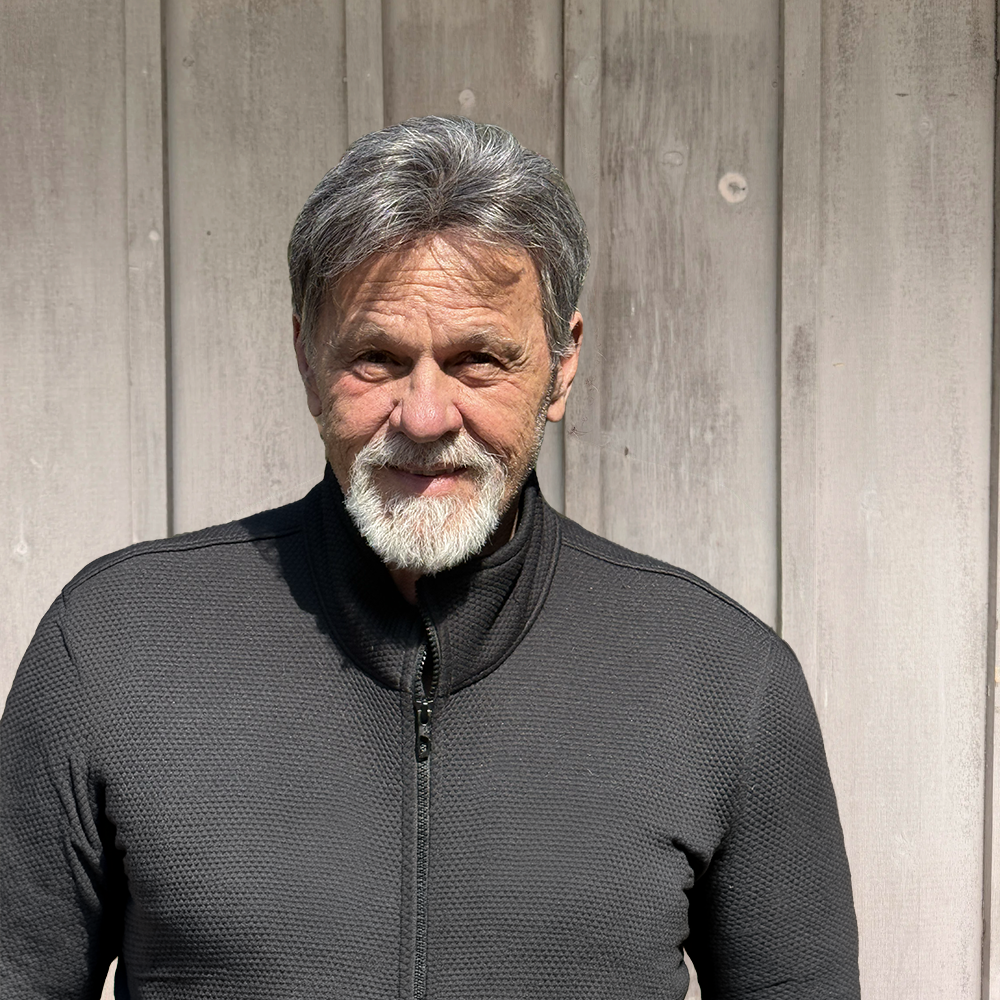 A smiling elderly man with gray hair and a beard, wearing a black zip-up jacket, stands outdoors in front of a wooden fence.