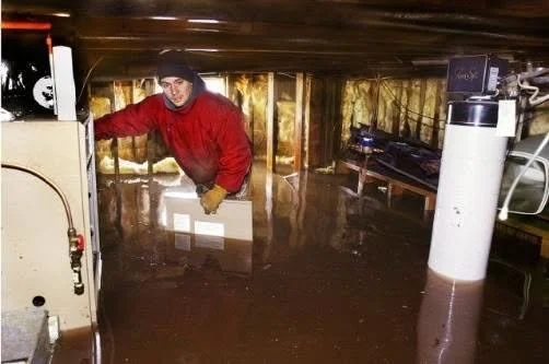 A person standing in a flooded basement, wearing a red jacket and black beanie, surrounded by water that covers the floor and some equipment.