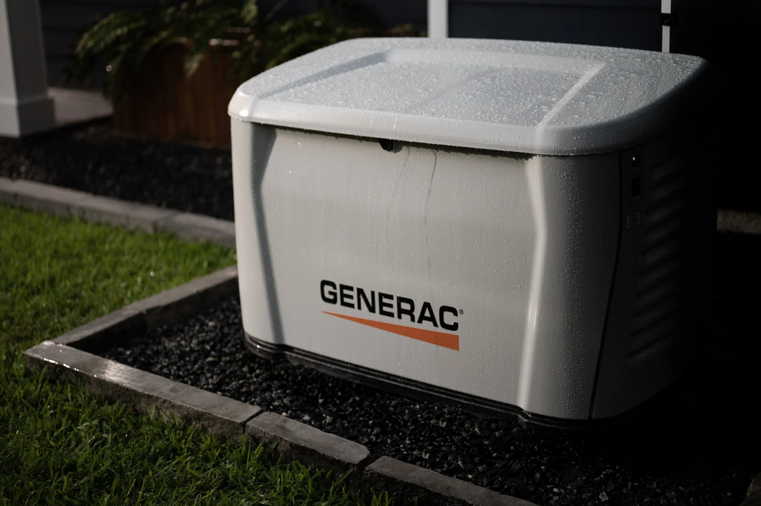 A Generac home standby generator installed outdoors with a sprinkler system, surrounded by grass and landscaping, during evening.