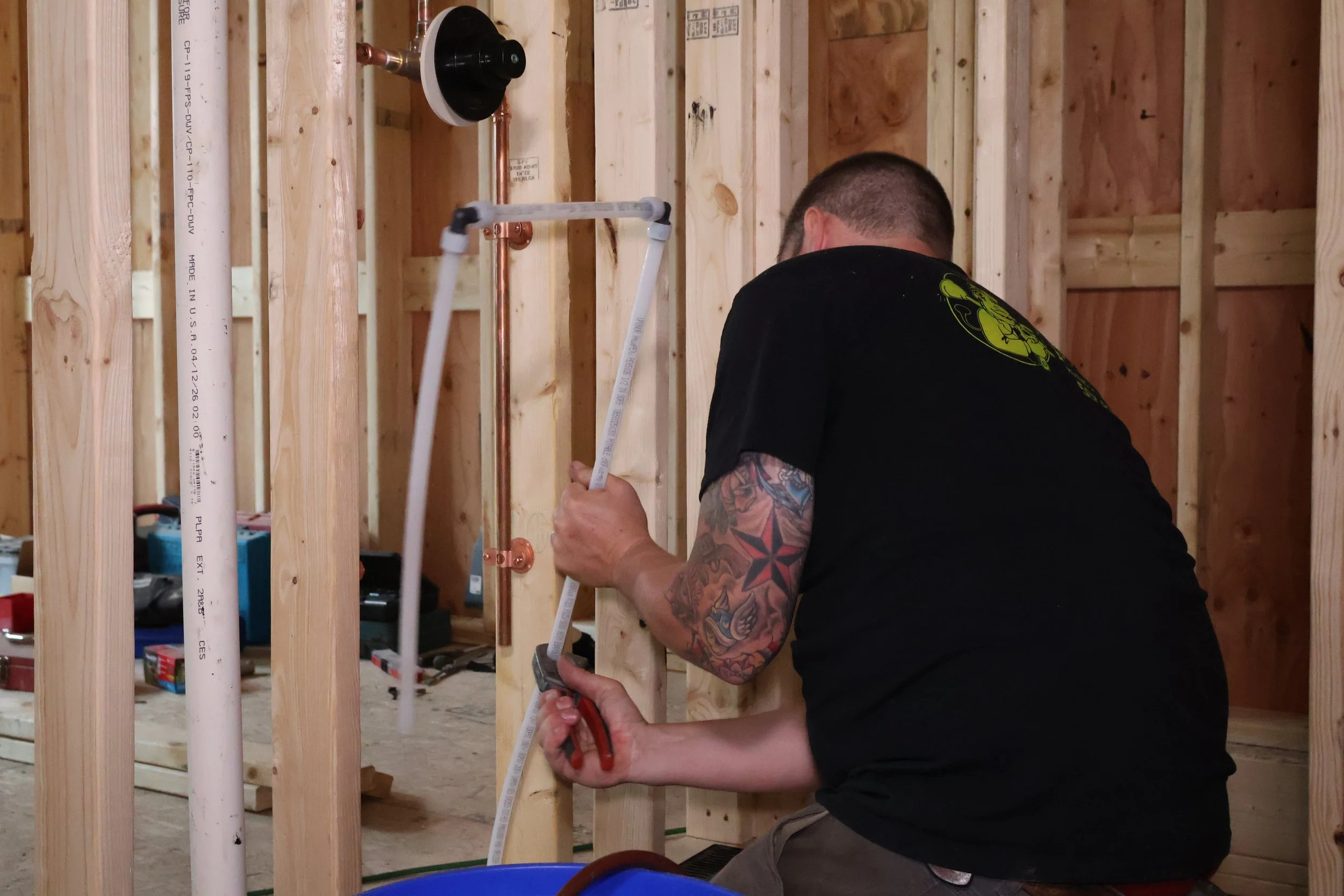 A man with tattoos on his arms working on plumbing inside a wooden framed wall during construction.