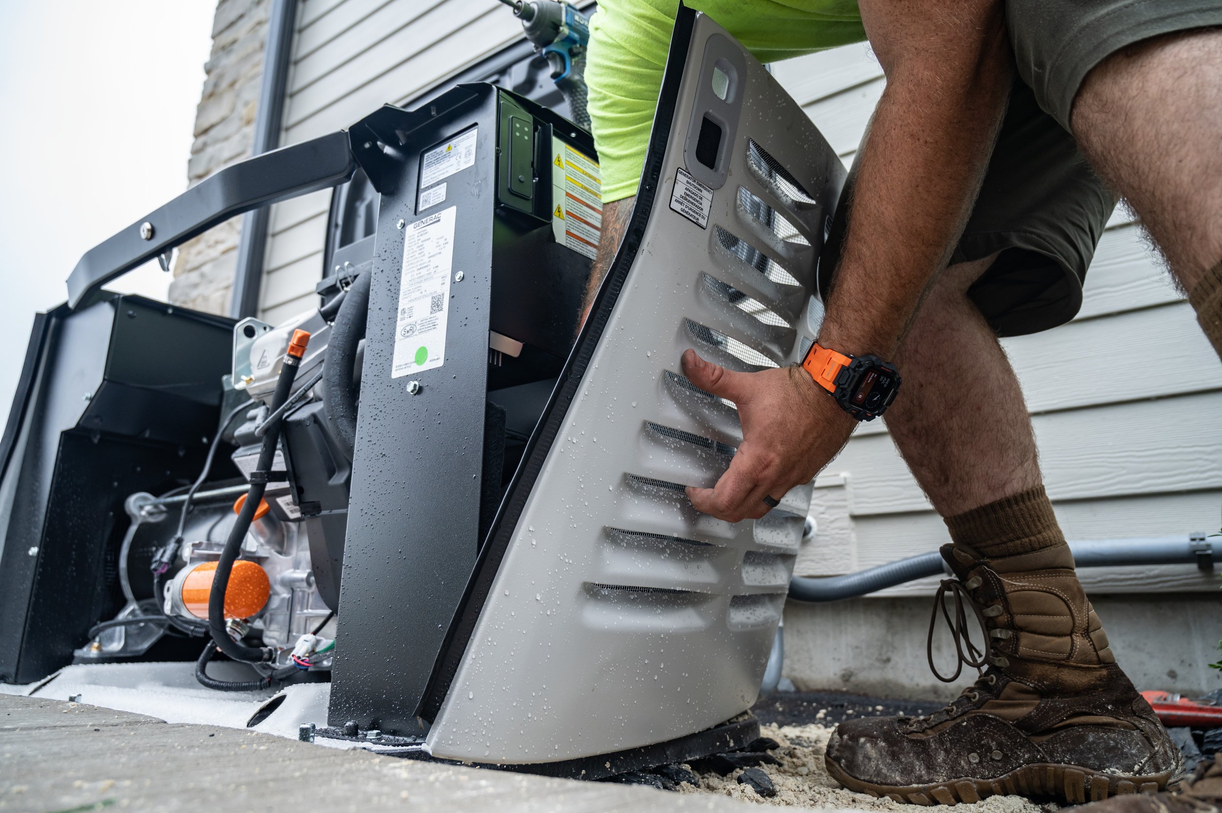 Person installing or repairing an HVAC unit outside a house, kneeling on the ground and holding a metal panel, with tools and equipment nearby.