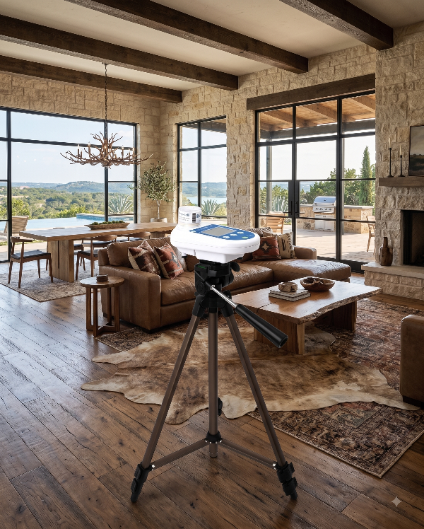 Living room with large windows, wooden beams, stone fireplace, beige sofa with pillows, wooden coffee table, and a device on a tripod in the center.