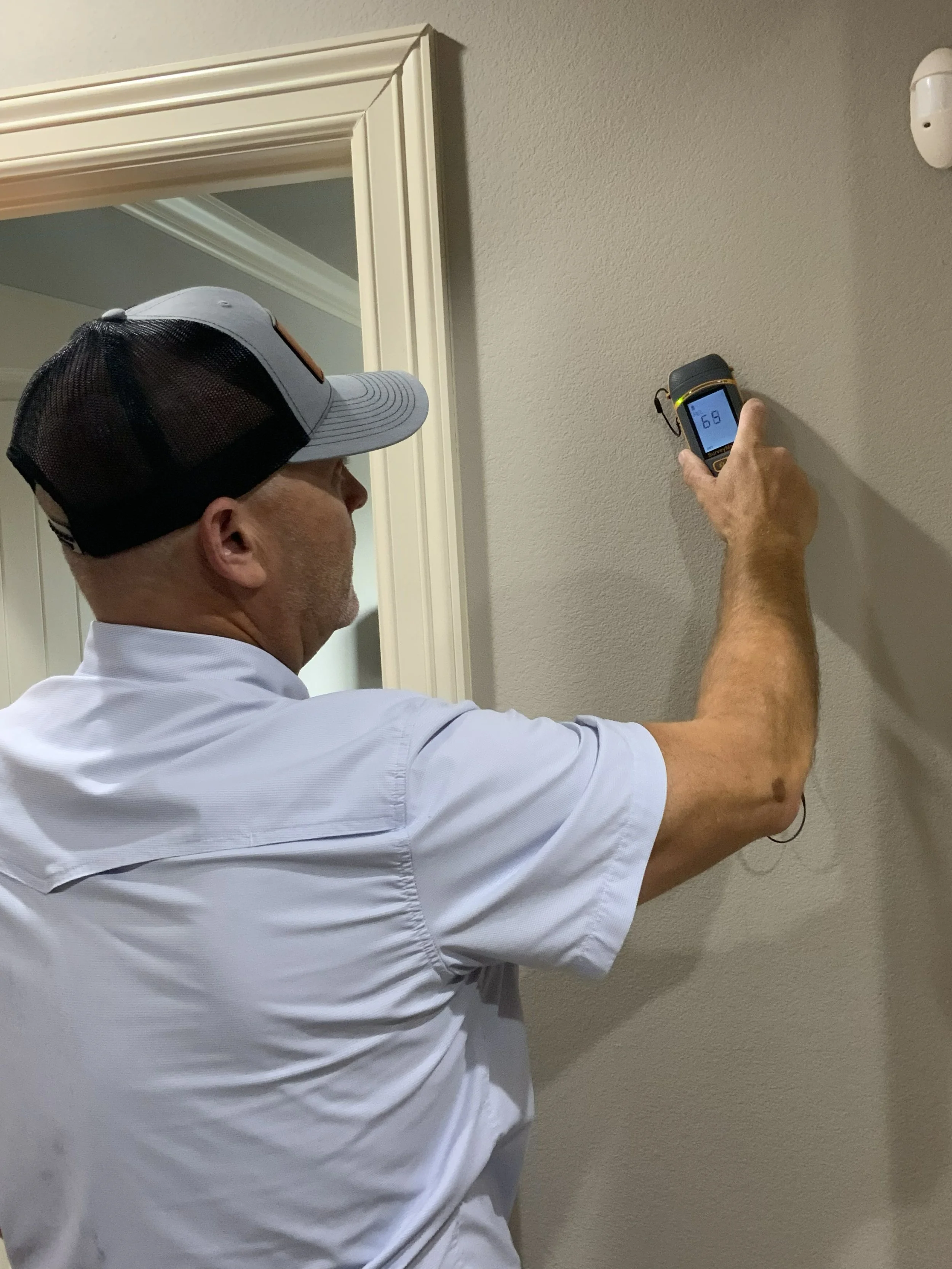 Michael Herbst, pricinciple consultant with ATX Mold Solutions with a gray and black trucker hat and a white shirt, using a moisture meter to measure moisture levels of drywall as part of a mold assessment