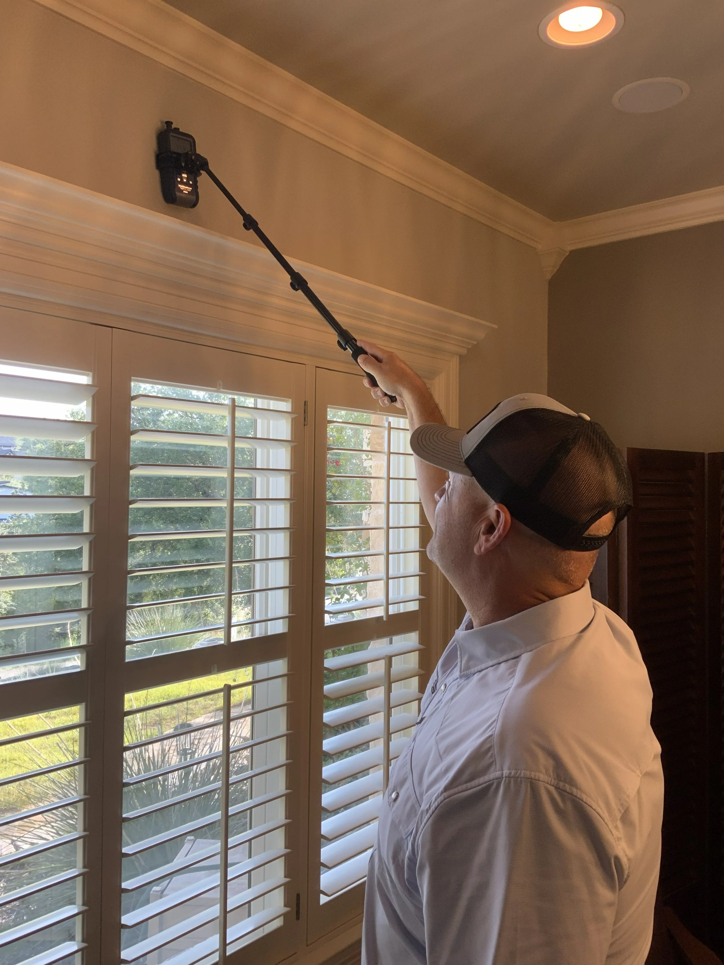 A man wearing a white shirt and a black cap is using a selfie stick to take a photo or video near a window with white plantation shutters. The interior has beige walls and a ceiling with recessed lighting.