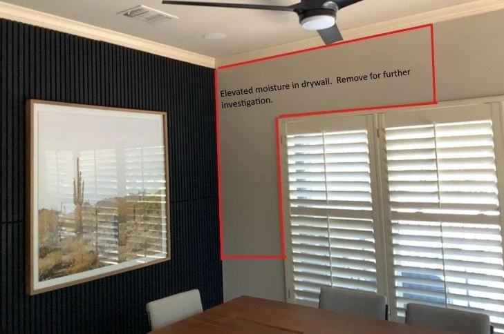 A dining room with dark wall paneling, two large windows with white shutters, a wooden table, and a ceiling fan. A red outline highlights a section of drywall with a note about elevated moisture, indicating the need for further investigation.