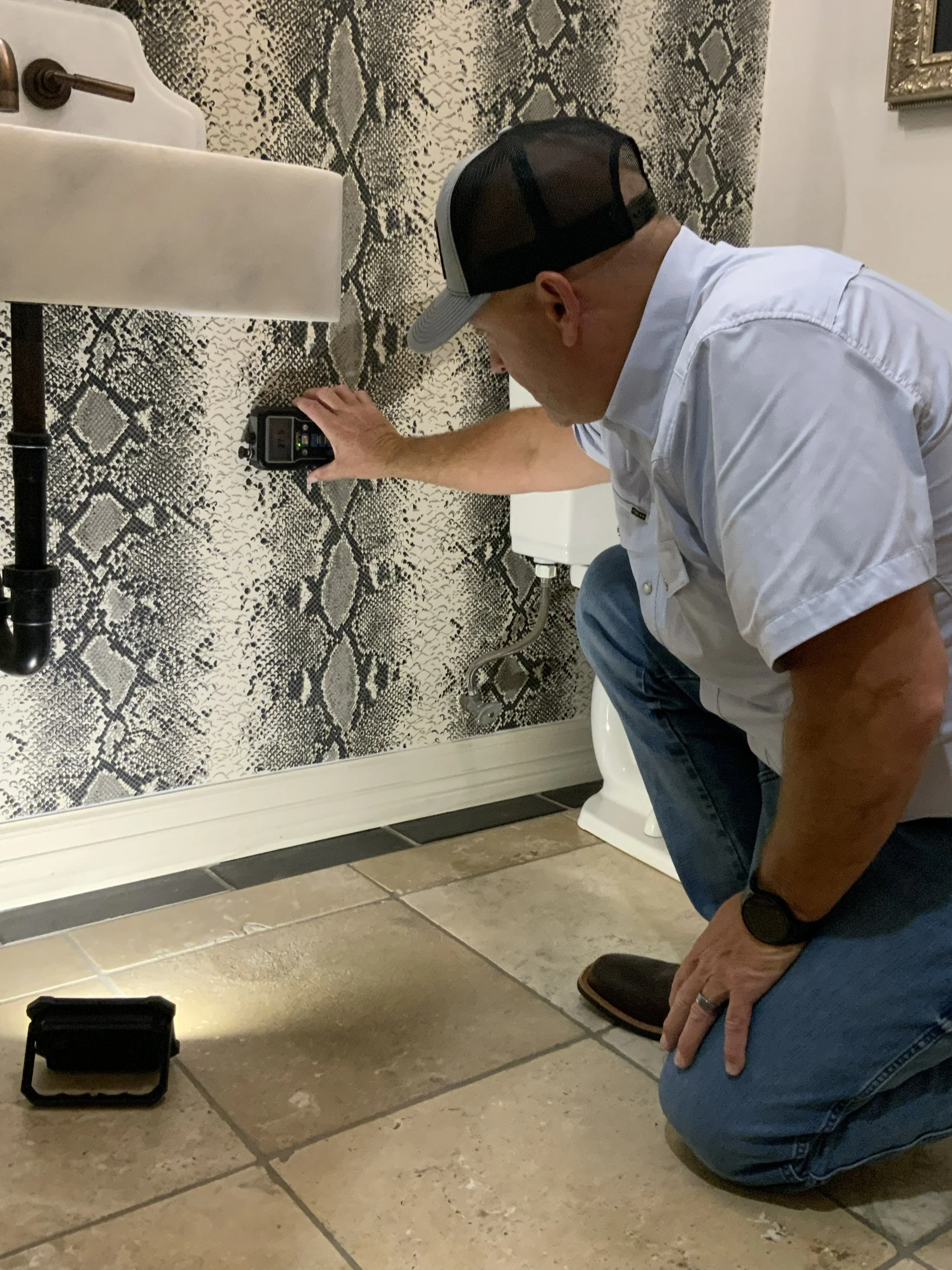 A man wearing a white shirt, blue jeans, and a mesh trucker hat kneels on the floor in a bathroom while using a sound level meter placed on the floor. The bathroom has a snake skin-patterned wall, a white toilet, and a black pipe under the sink.