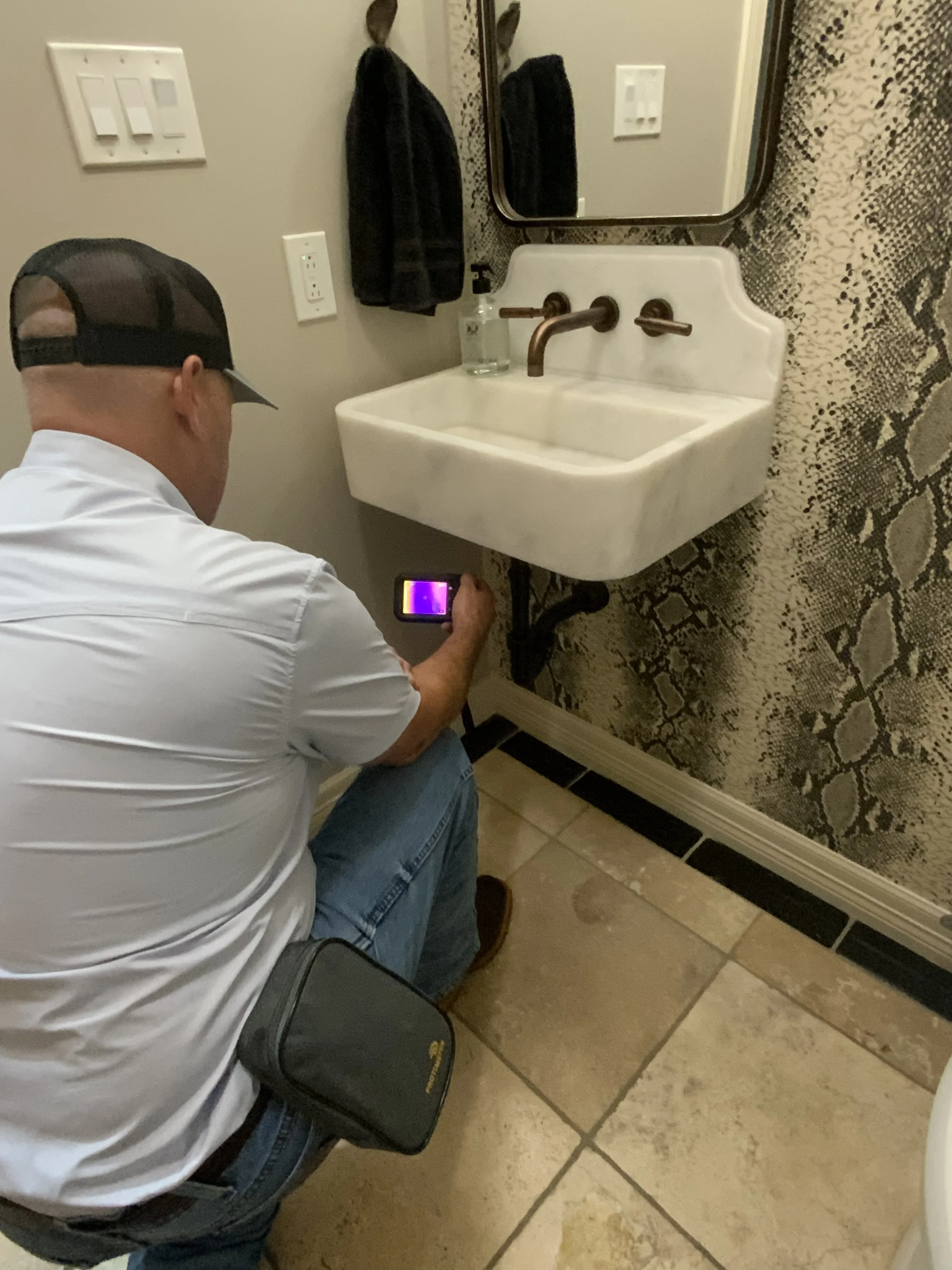 A man in a baseball cap and white shirt sits on a chair in front of a bathroom vanity with a mirror, sink, and soap dispenser, holding a device with a thermal camera displaying a colorful thermal image.