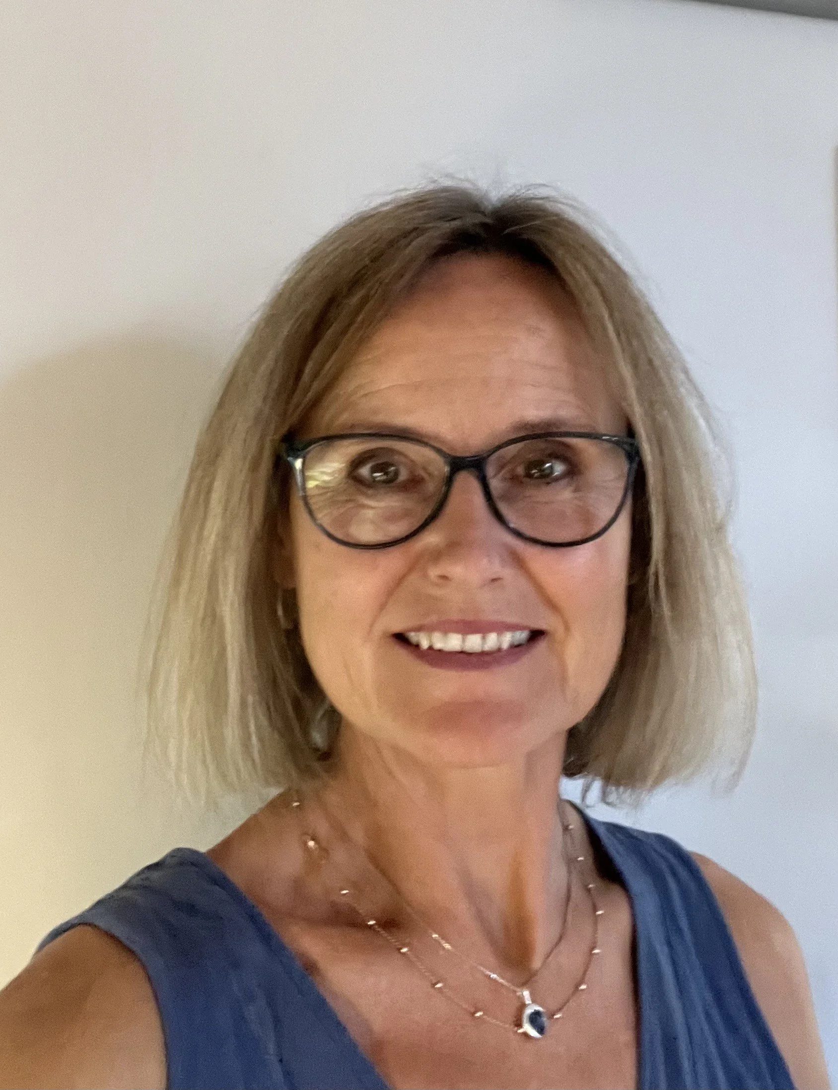 A middle-aged woman with blonde hair wearing glasses, a blue sleeveless top, and a necklace, smiling in front of a plain wall.