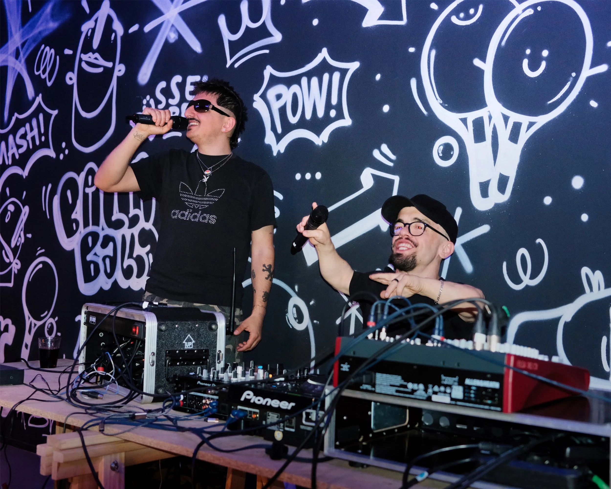 Zwei Männer, die auf einer Tanzfläche mit einer DJ-Ausrüstung auf einem Tisch stehen, während sie singen und lachen. Im Hintergrund eine Wand mit Kunstzeichnungen und Texten wie "WOW!", "SMASH!" und "POW!"