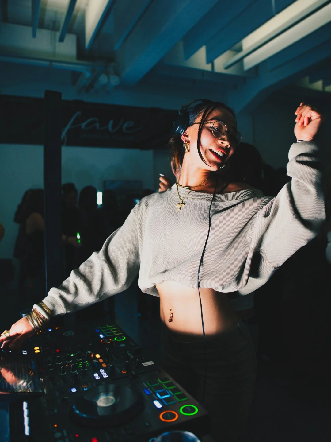 Eine smiling DJ-Dame mit Kopfhörern, Sonnenbrille und Piercings, die auf einer Tanzfläche an einem Mischpult Musik auflegt, umgeben von tanzenden Menschen