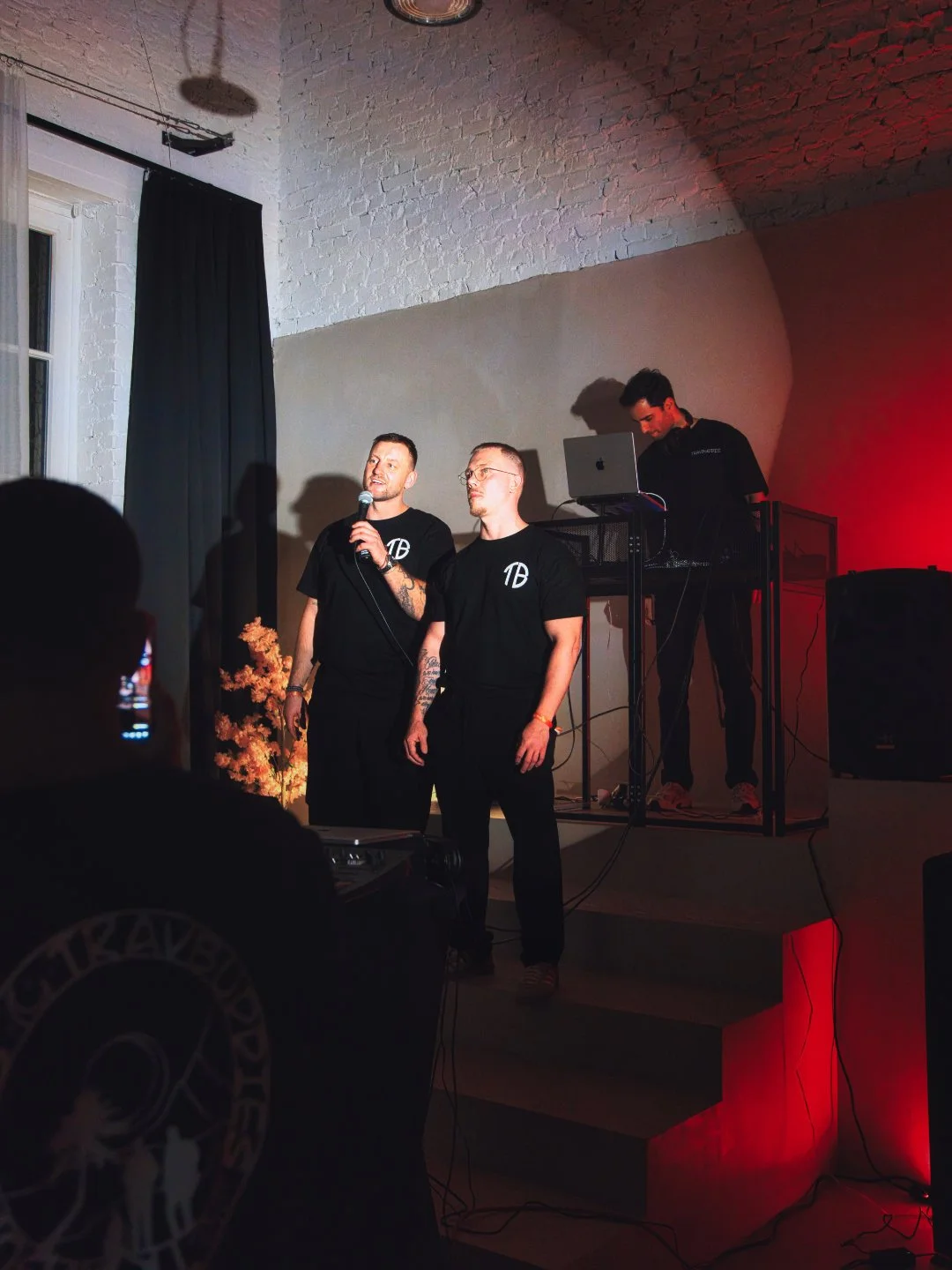 Zwei Männer stehen auf einer Bühne, einer spricht in ein Mikrofon, während im Hintergrund ein DJ an einem Laptop arbeitet