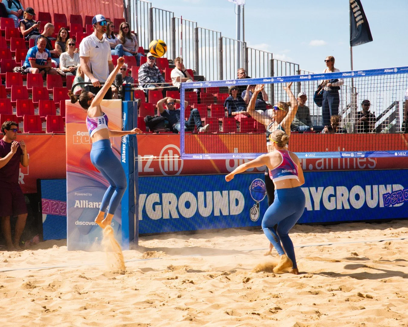 Zwei Beachvolleyballspielerinnen springen in der Luft, um den Ball über das Netz zu schlagen, während das Publikum im Hintergrund zusieht