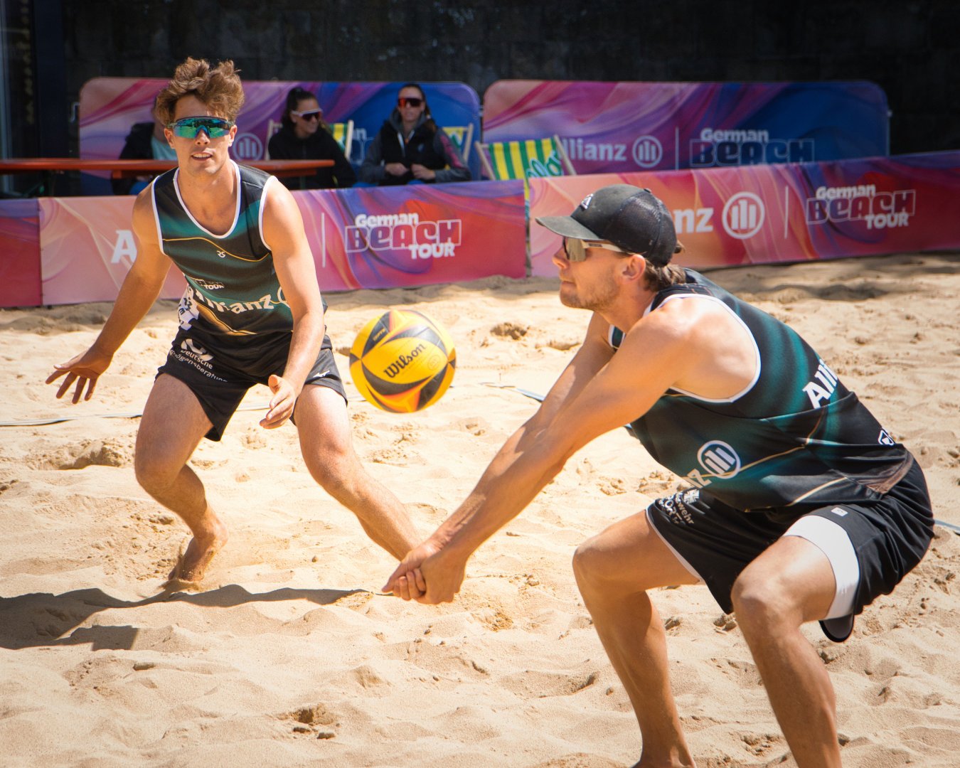 Zwei Beach-Volleyballspieler beim Spiel auf Sand, einer passt den Ball, während der andere sich für einen Distanzsprung vorbereitet, im Hintergrund sind Zuschauer und bunte Werbung