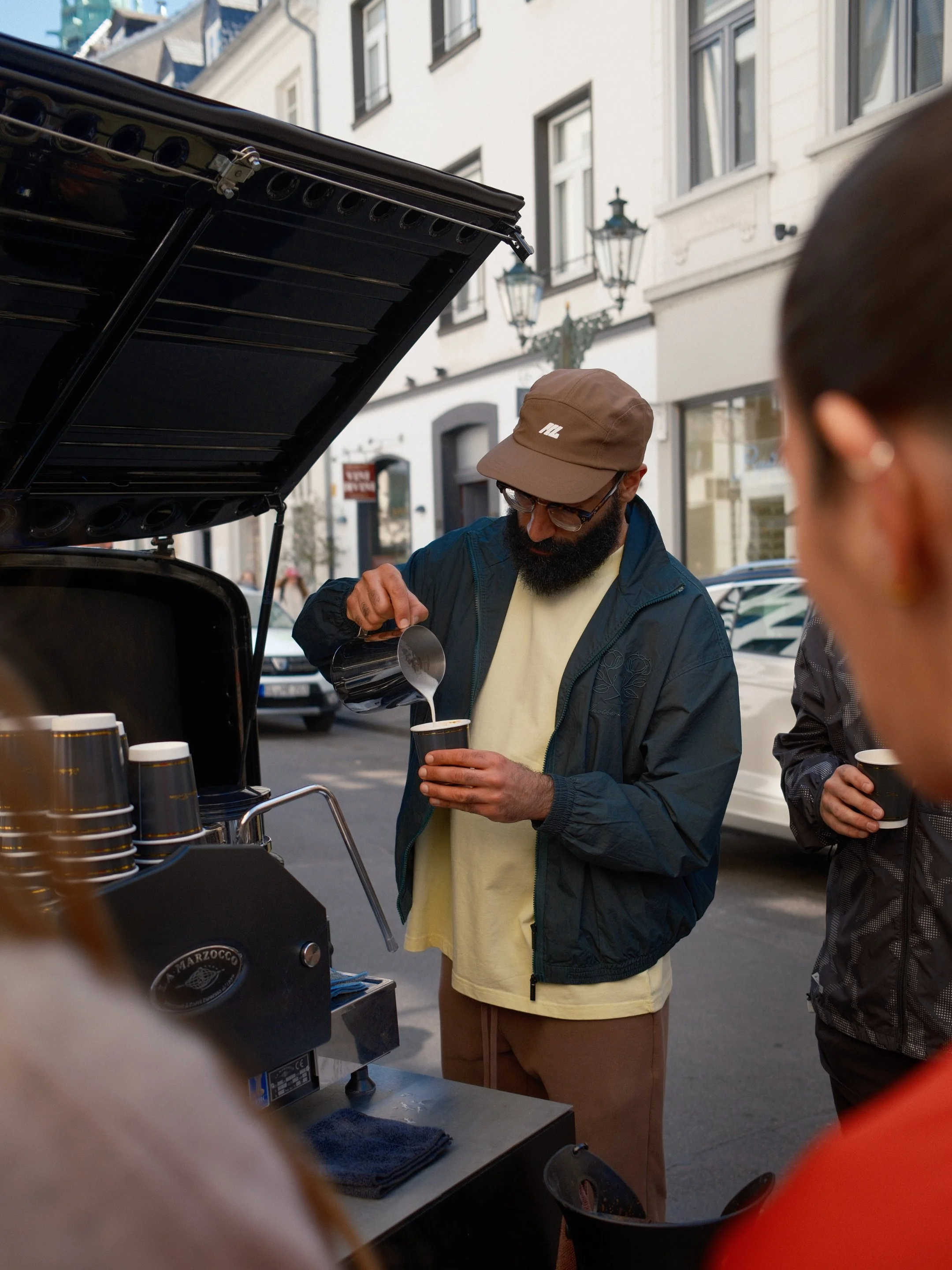 Mann mit Brille und Mütze, der Kaffee aus einem Espressomaschinenbehälter in eine Pappbecher füllt, während eine andere Person daneben steht, auf einer Straße mit alten Gebäuden im Hintergrund