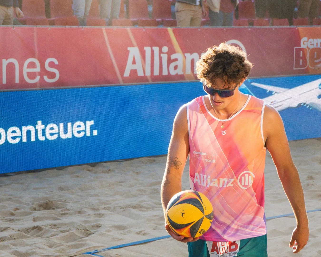 Beachvolleyballspieler mit Sonnenbrille hält einen Ball auf Sandplatz, im Hintergrund Werbebanner und Zuschauer