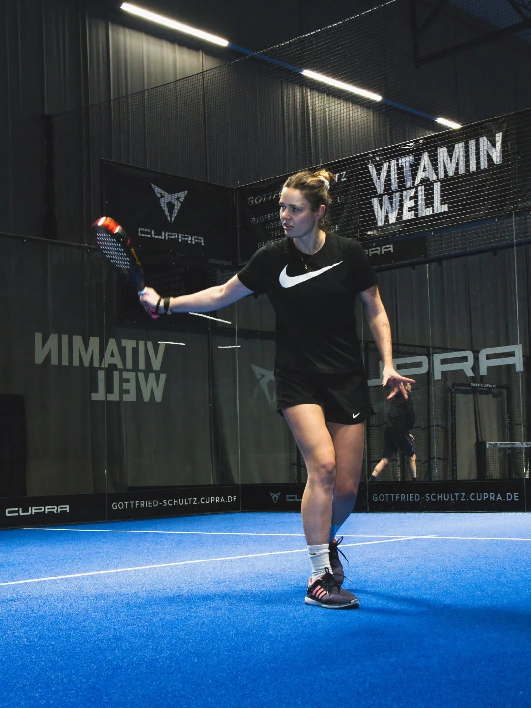Eine junge Frau spielt Padel in einer Halle auf blauem Boden, trägt schwarze Sportkleidung mit Nike-Logo, hält einen Tennisschläger und wird beim Rückschlag beobachtet
