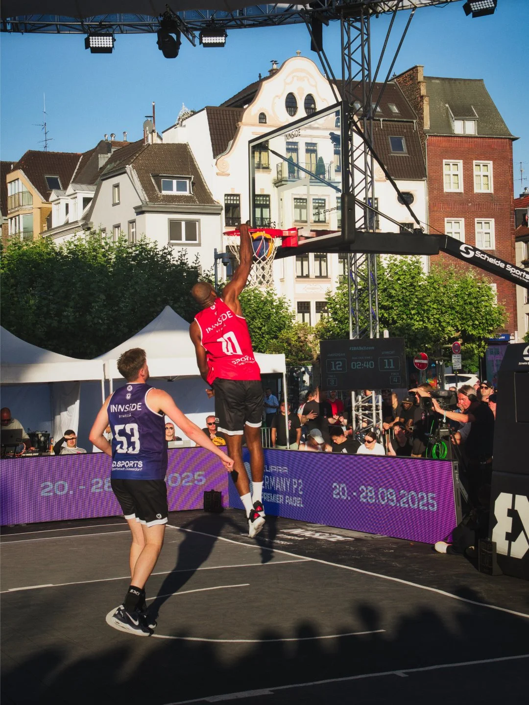 Zwei Basketballspieler beim Dunking während eines Spiels im Freien auf einem Stadtplatz, im Hintergrund historische Gebäude, Zuschauer und Kameras