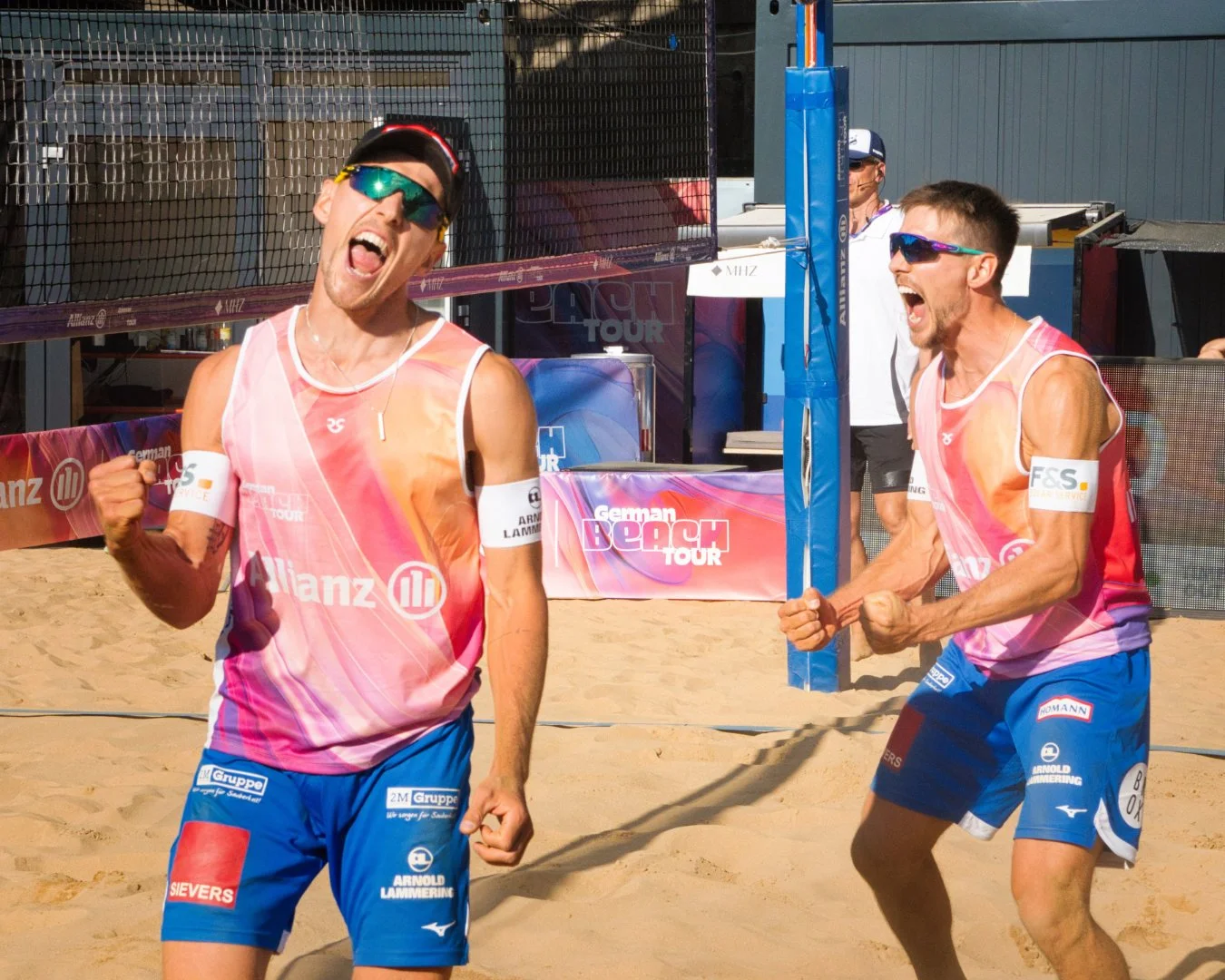 Zwei beachvolleyballspieler jubeln auf Sand, feiern einen Punkt während eines Spiels, tragen Sonnenbrillen und Sportkleidung mit Logos, im Hintergrund ein Netz und eine Zuschauertribüne