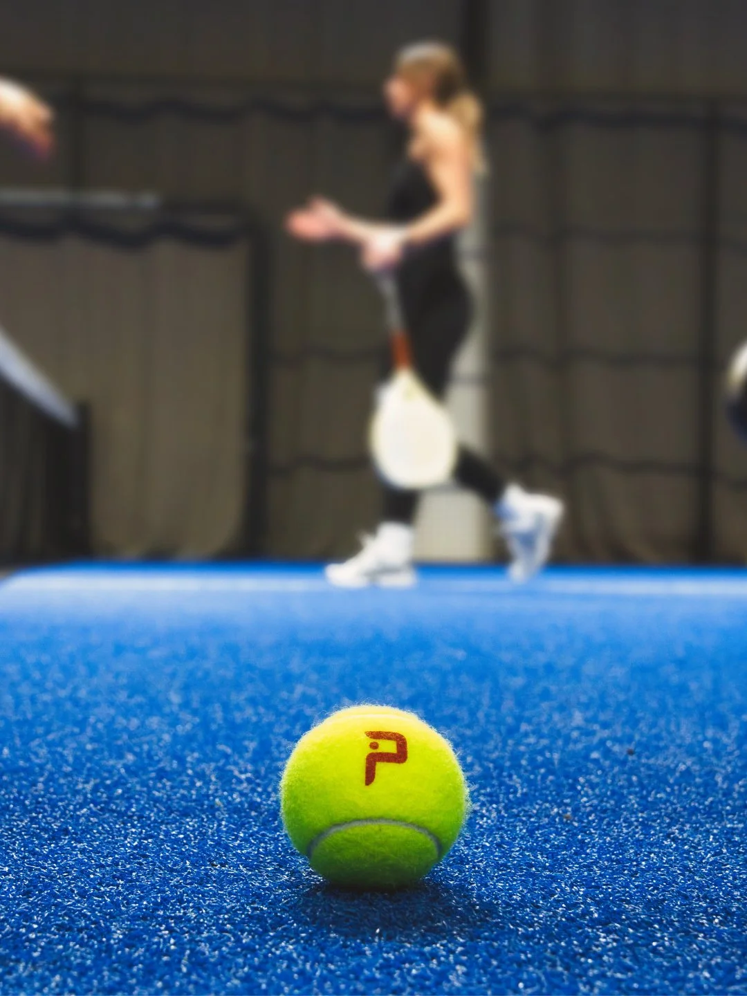 Gelber Tennisball mit rotem Logo auf blauer Tennisplatz im Hintergrund eine Tennisspielerin mit Tennisschläger in Aktion