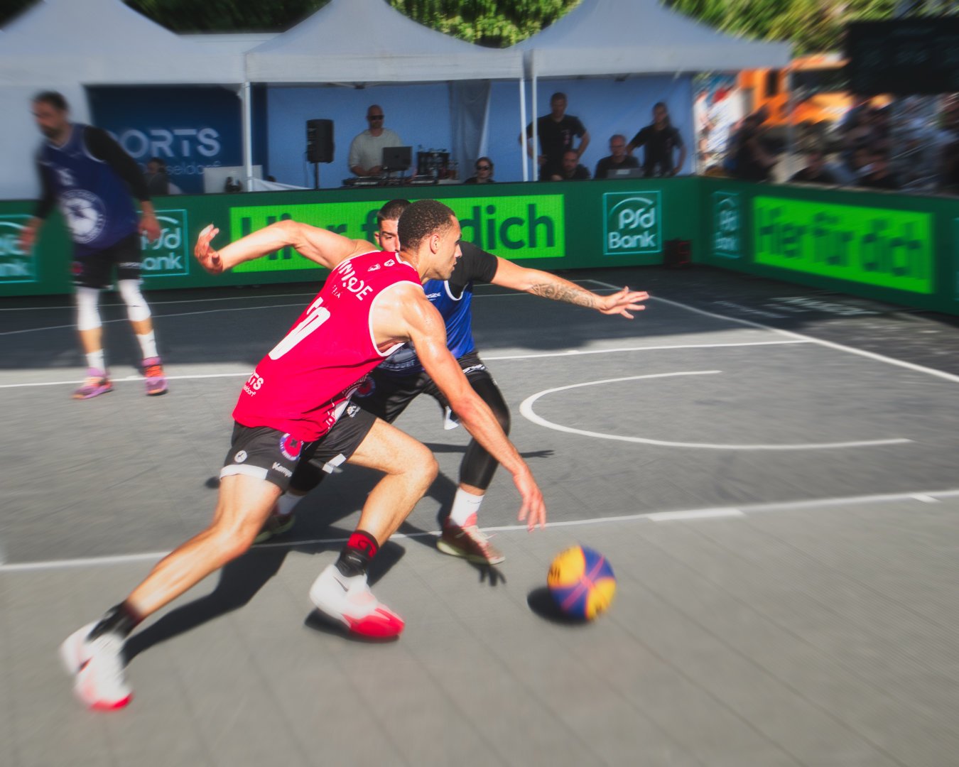 Zwei Basketballspieler verfolgen den Ball während eines Spiels auf einem Freiplatz, im Hintergrund sind Zuschauer und eine Unterstand mit Lautsprechern
