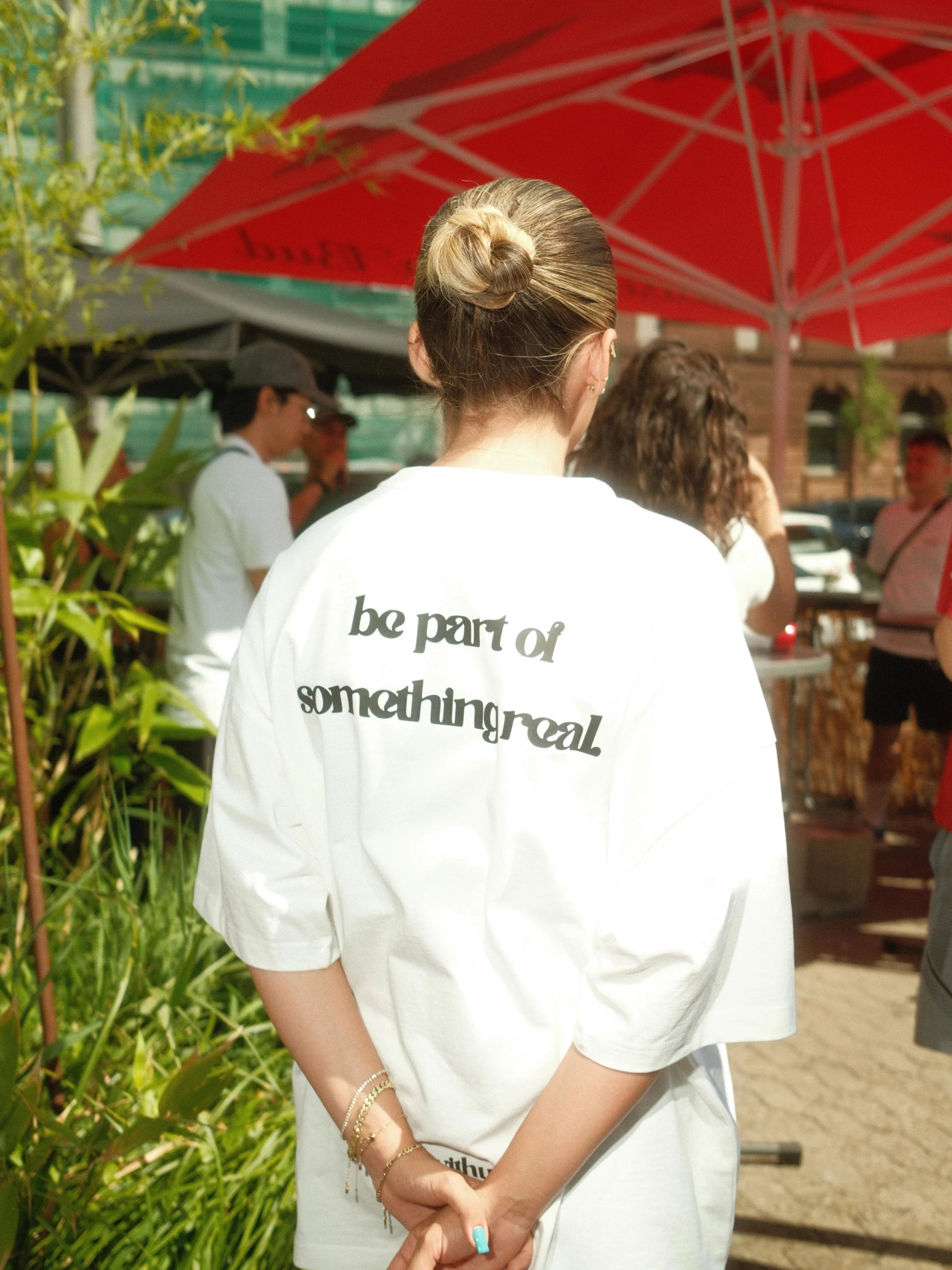 Eine Frau trägt ein weißes T-Shirt mit der Aufschrift 'be part of something real', während sie an einem sonnigen Tag im Freien steht, umgeben von Pflanzen und einem roten Sonnenschirm im Hintergrund