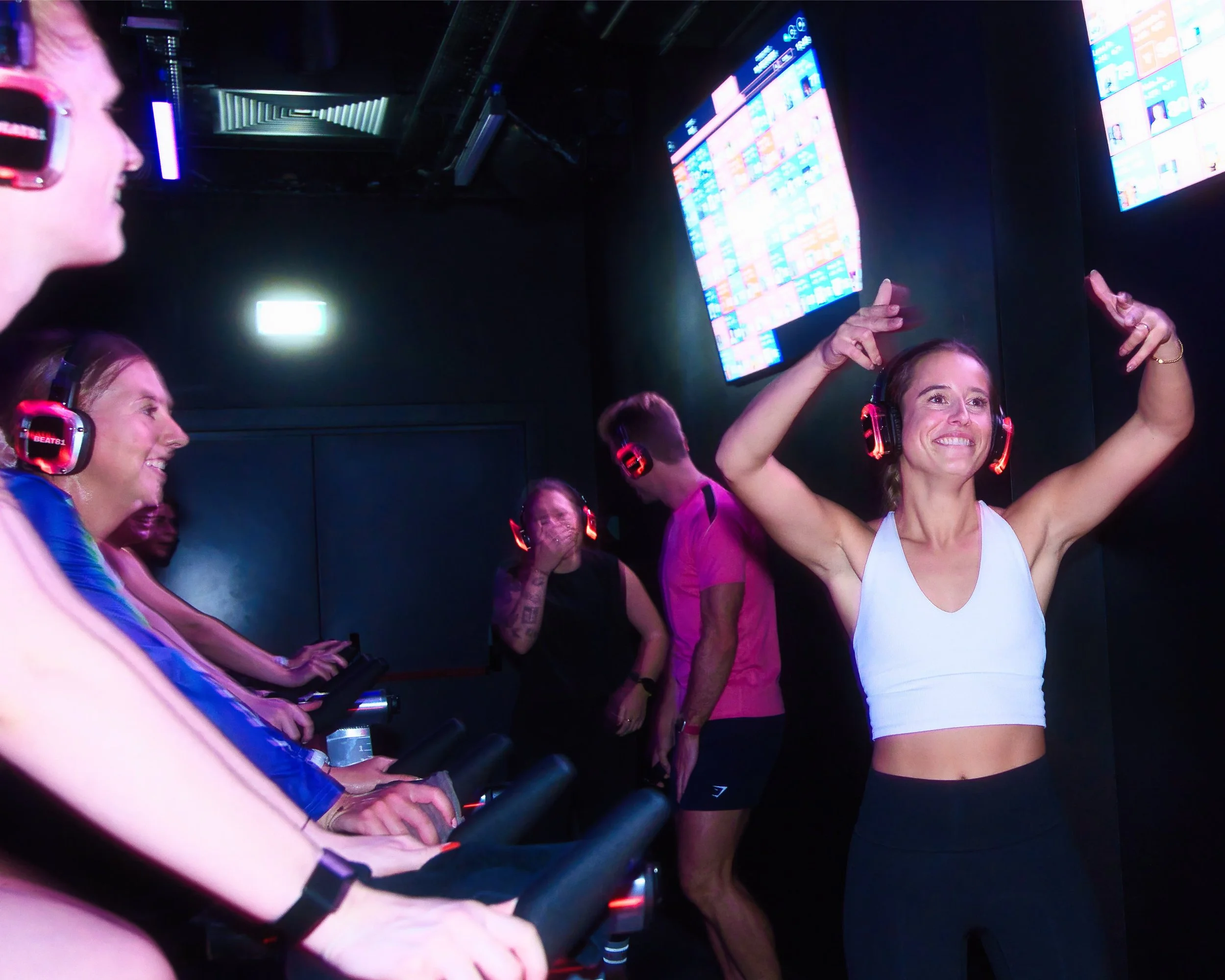 Mehrere Frauen im Fitnessstudio mit Kopfhörern, die auf Laufbändern laufen, eine Frau in einem weißen Sporttop hebt die Arme und lächelt