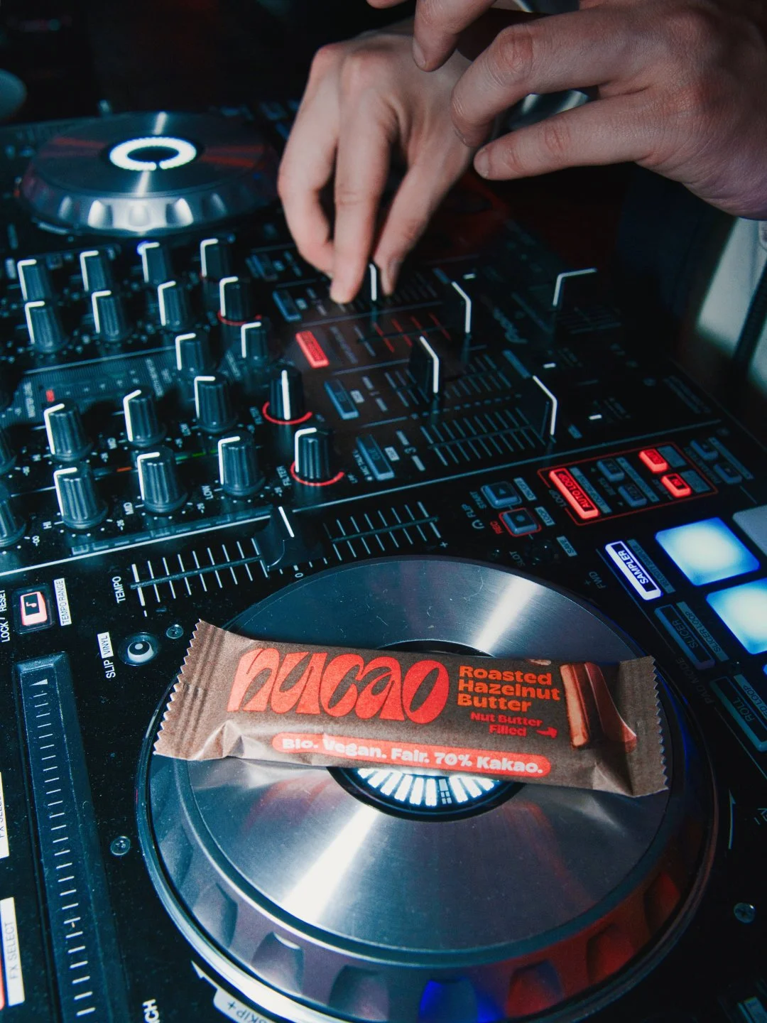 Ein DJ-Controller mit leuchtendem Display, über dem ein Hand die Regler bedient, und eine Schokoladentafel mit der Aufschrift 'NUCO' und 'Roasted Hazelnut Butter' liegt auf dem Plattenteller