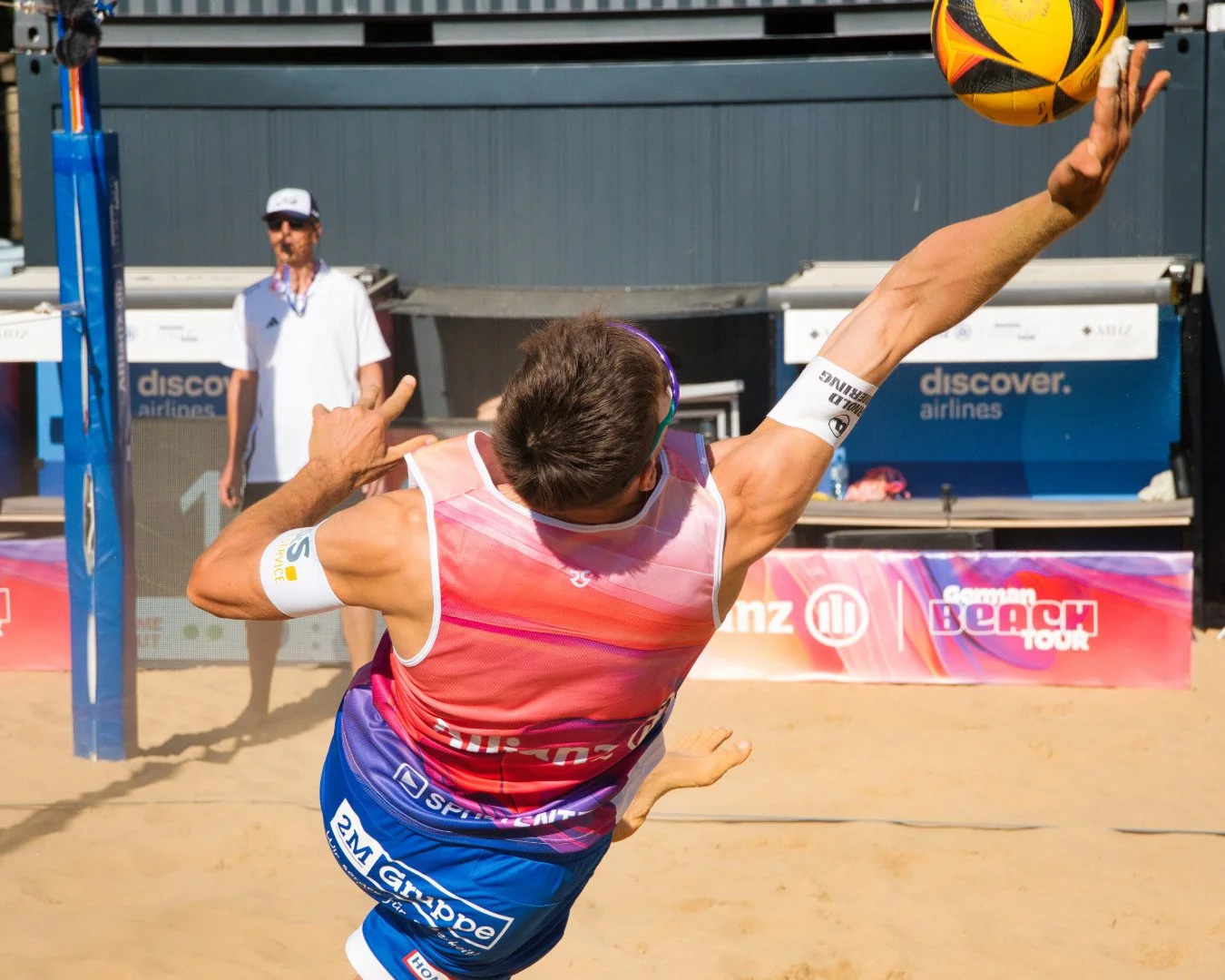 Ein männlicher Beachvolleyballspieler in einem pinken und blauen Trikot springt hoch, um den Ball im Sand zu schlagen. Im Hintergrund stehen Fotografen und Werbetafeln bei einem Beachvolleyballturnier