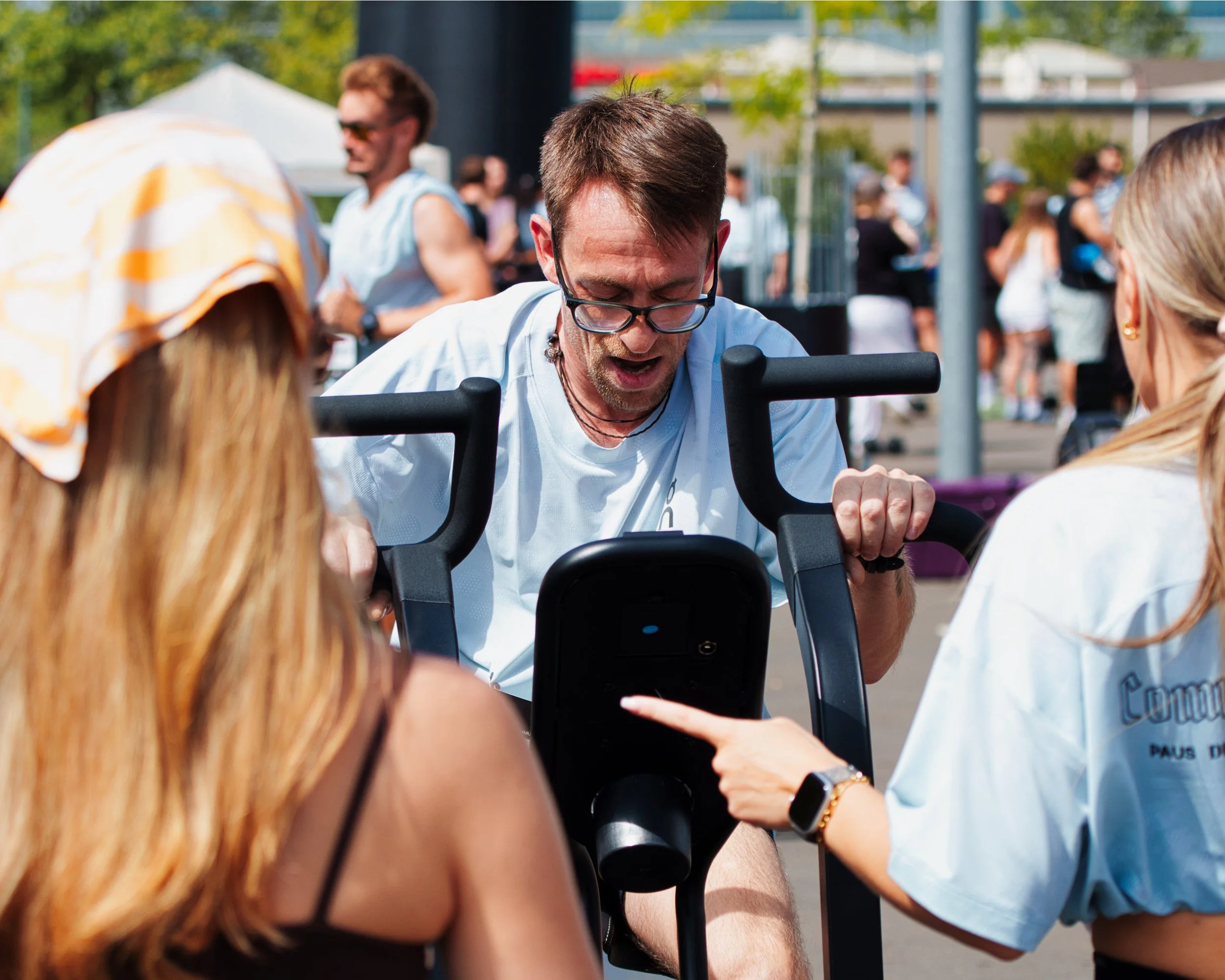 Eine Gruppe von Menschen im Freien, im Vordergrund ein Mann mit Brille, der auf einem Ergometer trainiert, umgeben von anderen Personen